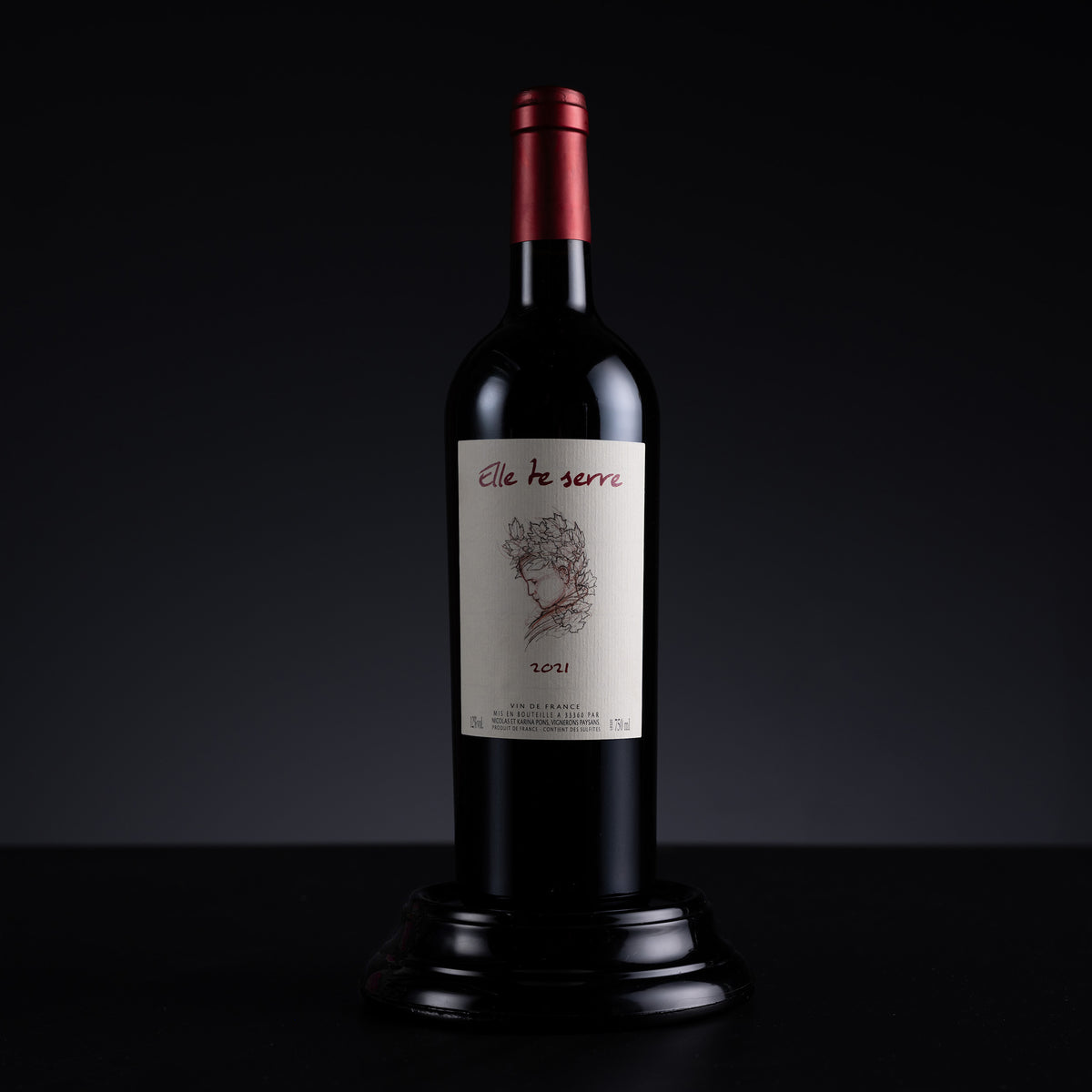 A bottle of red wine displayed on a black pedestal against a dark studio background. The bottle has a dark glass body, a metallic red foil top, and a cream-colored label with the brand name 'elle te serre' and a floral illustration.