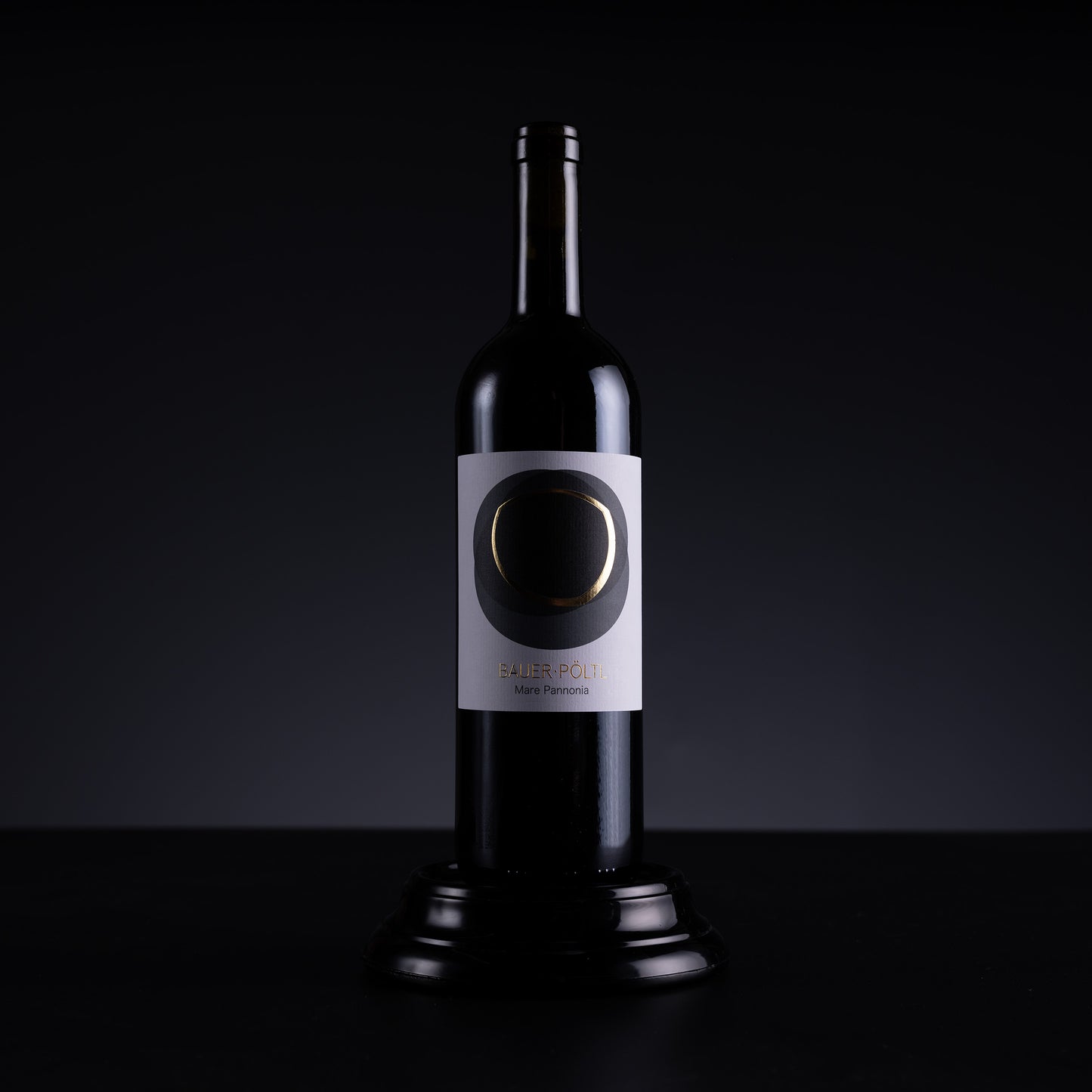 A dark glass wine bottle with a white label featuring a circular black and gold design, displayed on a black pedestal against a dark studio background.