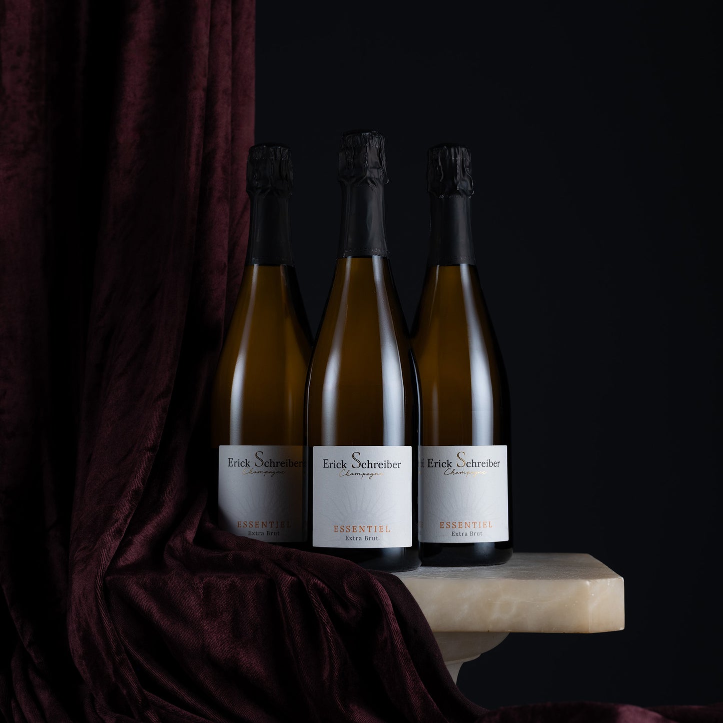 Three bottles of Erik Schreiber Essentia sparkling wine arranged on a light stone slab with a dark burgundy velvet drape against a black background.