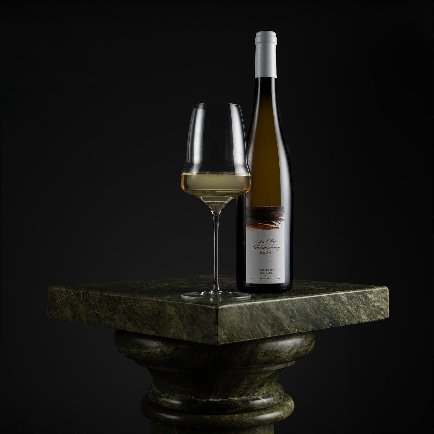 A bottle of white wine and a filled wine glass displayed on a dark green stone pedestal against a black background.