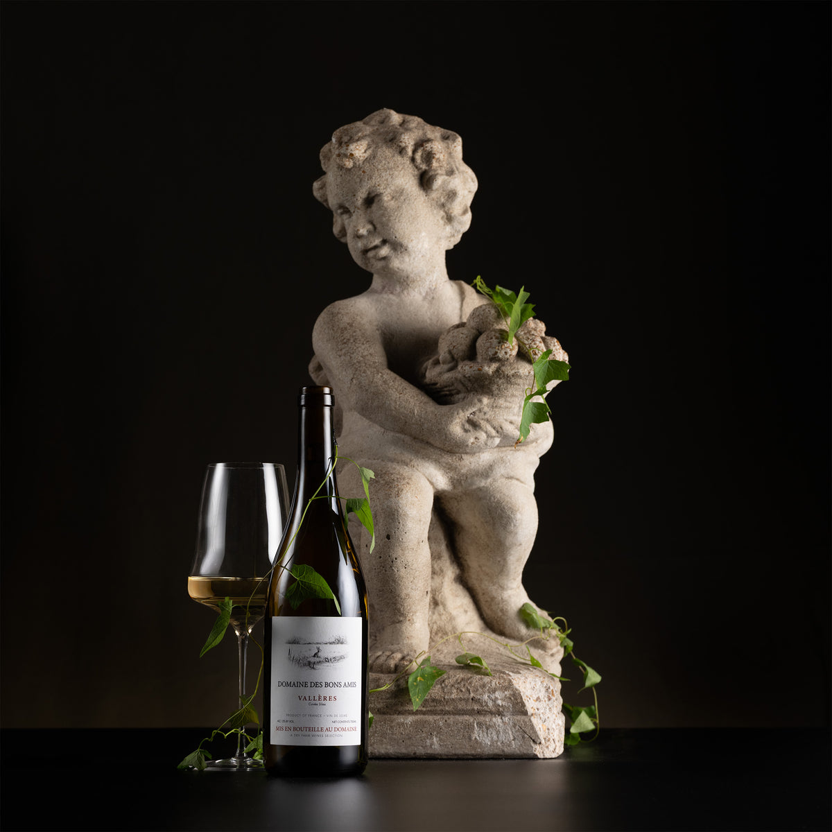 A wine bottle and a filled wine glass styled beside a small cherub statue holding a bouquet of green leaves, set against a dark black background.