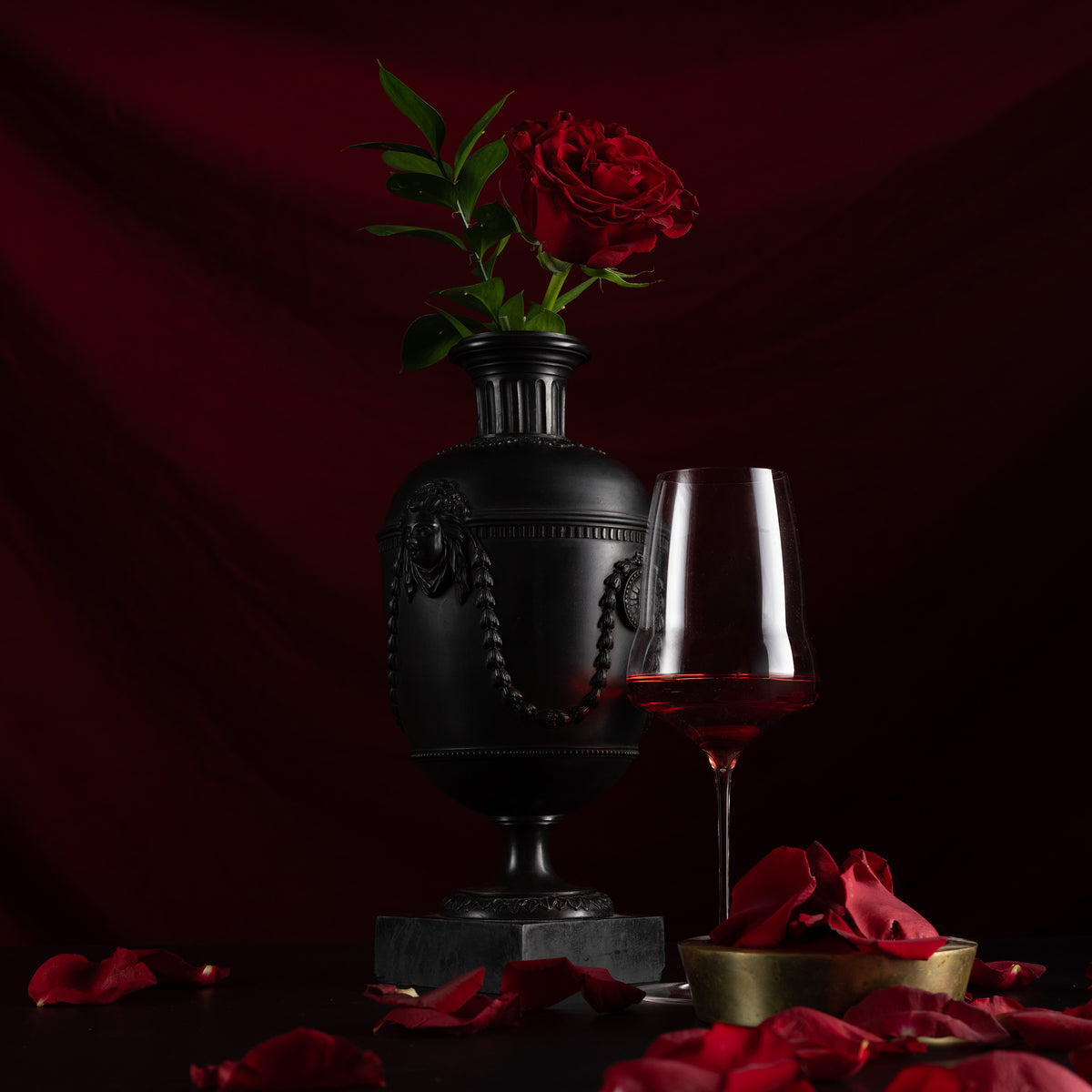 A black decorative urn-shaped vase holding a single red rose, styled beside a wine glass with red wine on a dark surface with scattered red petals against a deep red draped backdrop.