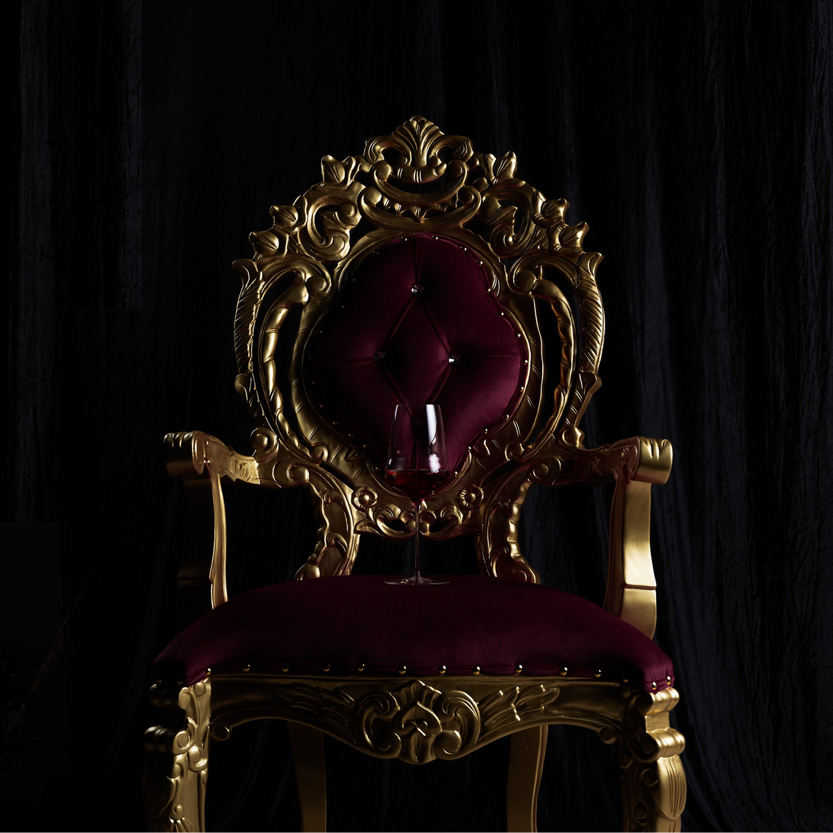 An ornate baroque-style throne chair with a carved gold frame and deep burgundy velvet upholstery, set against a dark black curtain background.