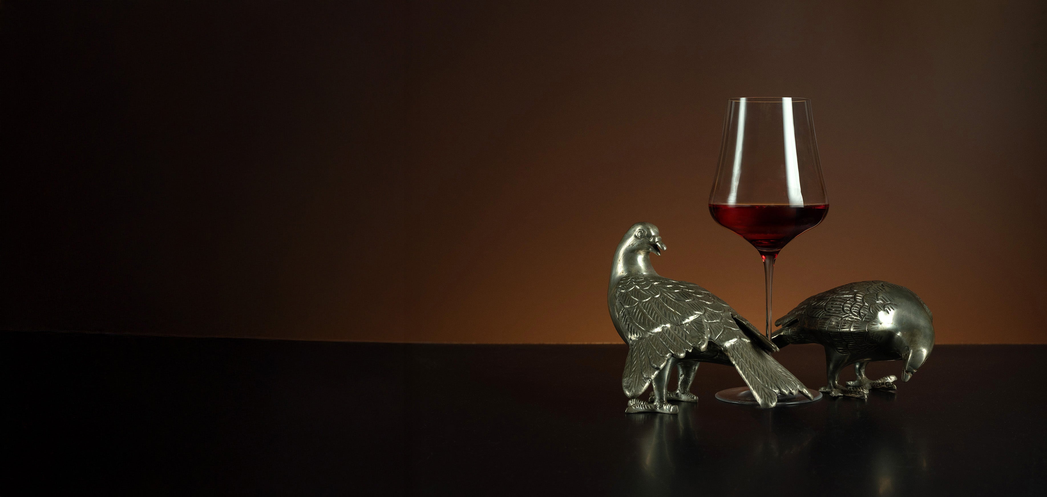 Two metallic animal figurines and a glass of red wine on a dark surface with a brown background