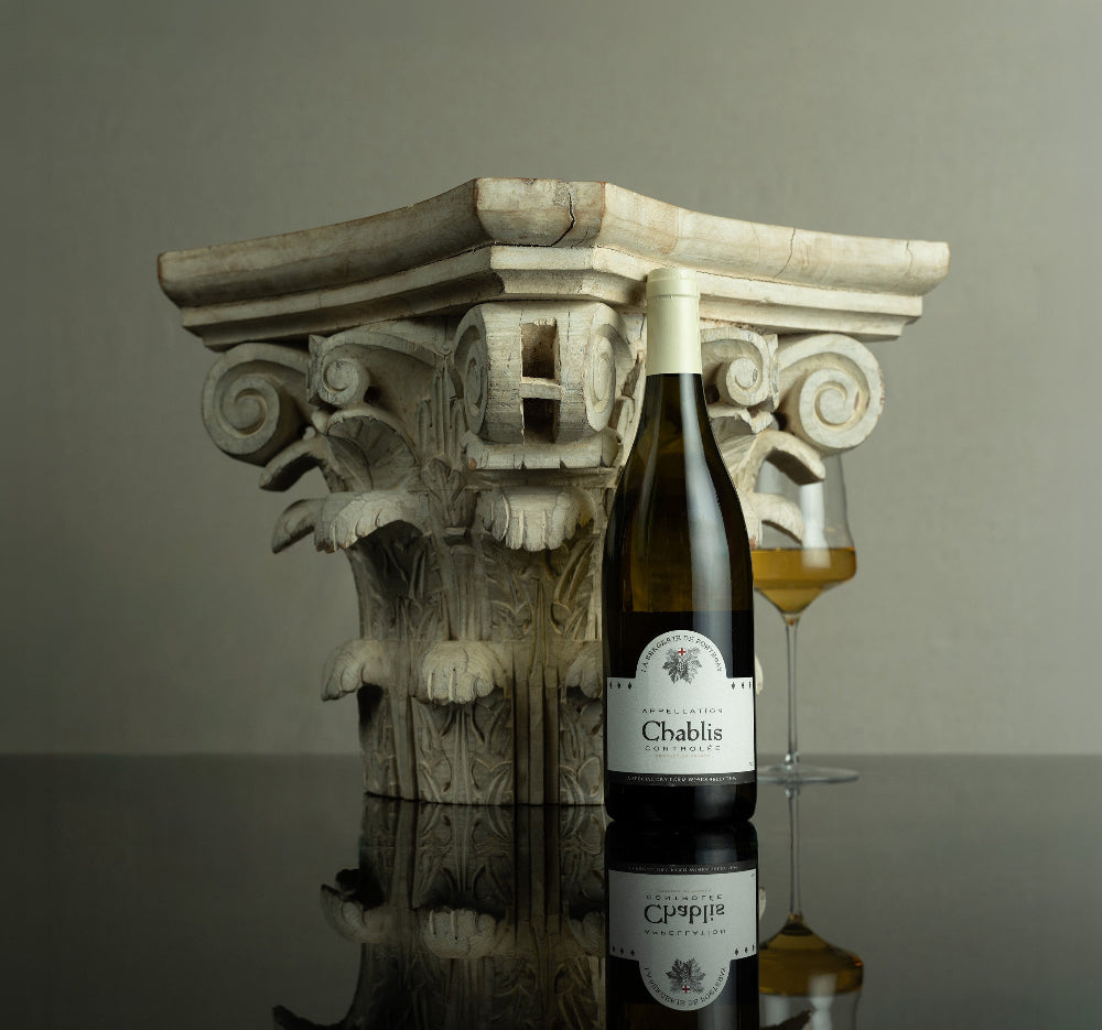 Wine bottle labeled 'Chablis' with a glass of wine on a reflective surface with an architectural column in the background.