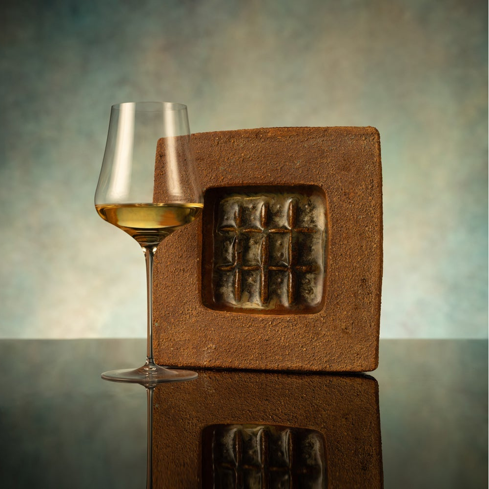 Glass of white wine next to a square wooden block with a grid pattern on a reflective surface.
