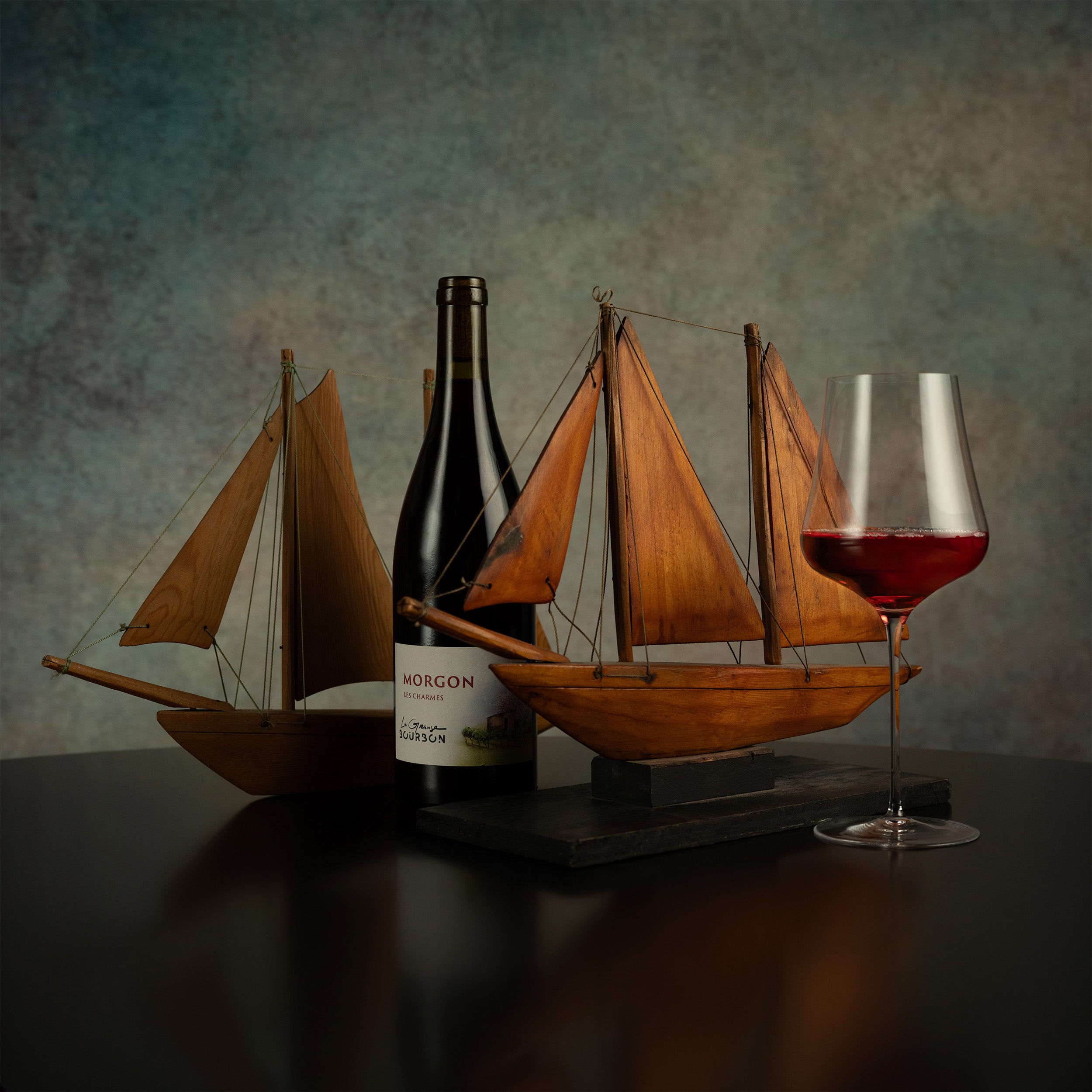 Bottle of wine with a wooden sailboat model and a glass of red wine on a dark surface.