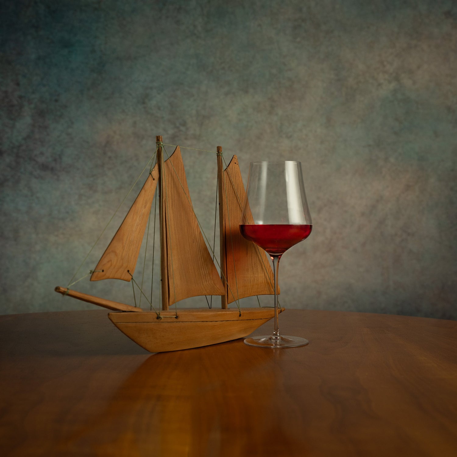 Wooden sailboat model and glass of red wine on a wooden surface with a textured wall background