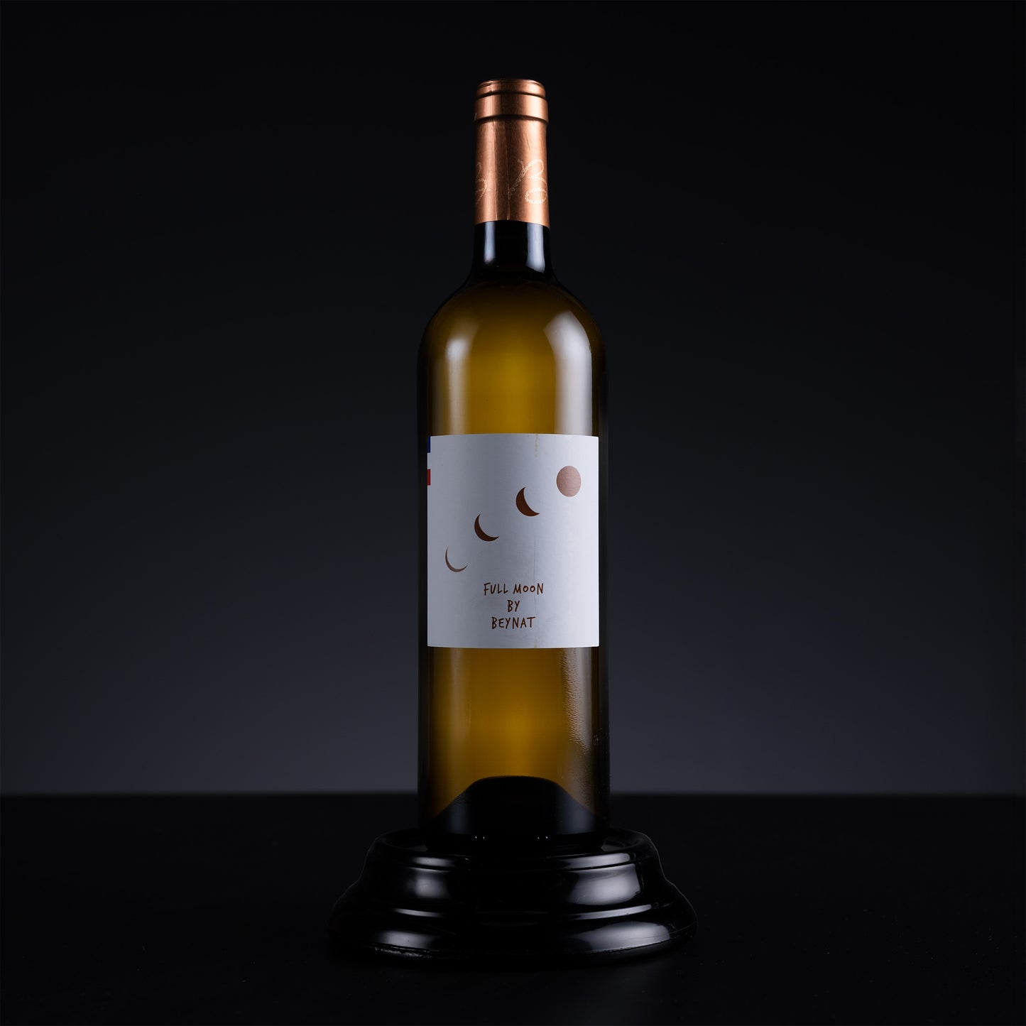 A single wine bottle with a white label featuring a minimalist moon and dot graphic, displayed on a black stand against a dark studio background.