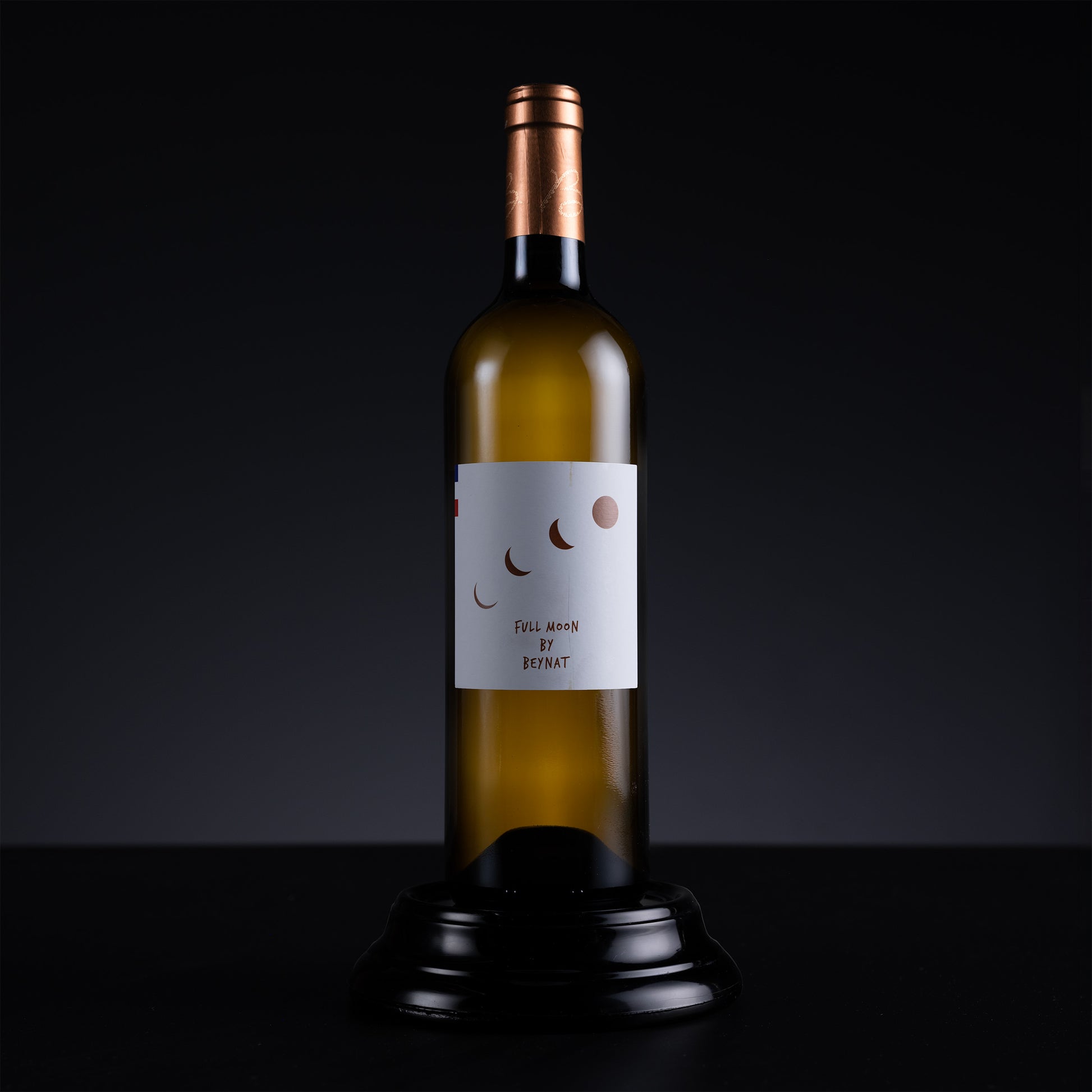 A single wine bottle with a white label featuring a minimalist moon and dot graphic, displayed on a black stand against a dark studio background.