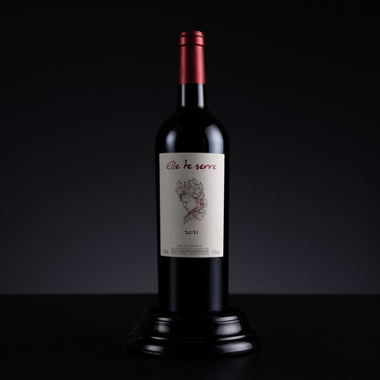 A bottle of red wine displayed on a black pedestal against a dark studio background. The bottle has a dark glass body, a metallic red foil top, and a cream-colored label with the brand name 'elle te serre' and a floral illustration.