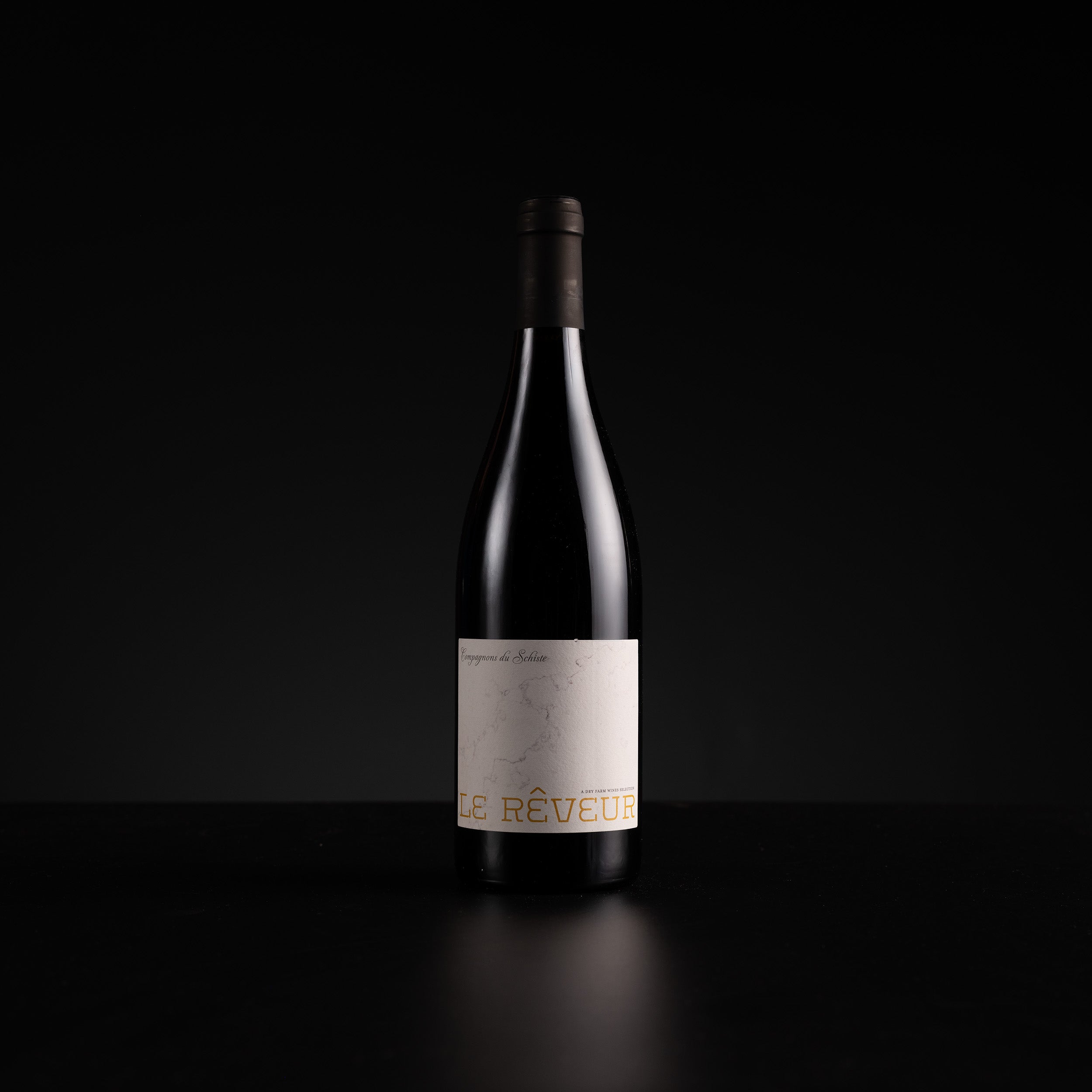 A single dark wine bottle with a minimalist off-white label and gold lettering, standing on a dark reflective surface against a black background.