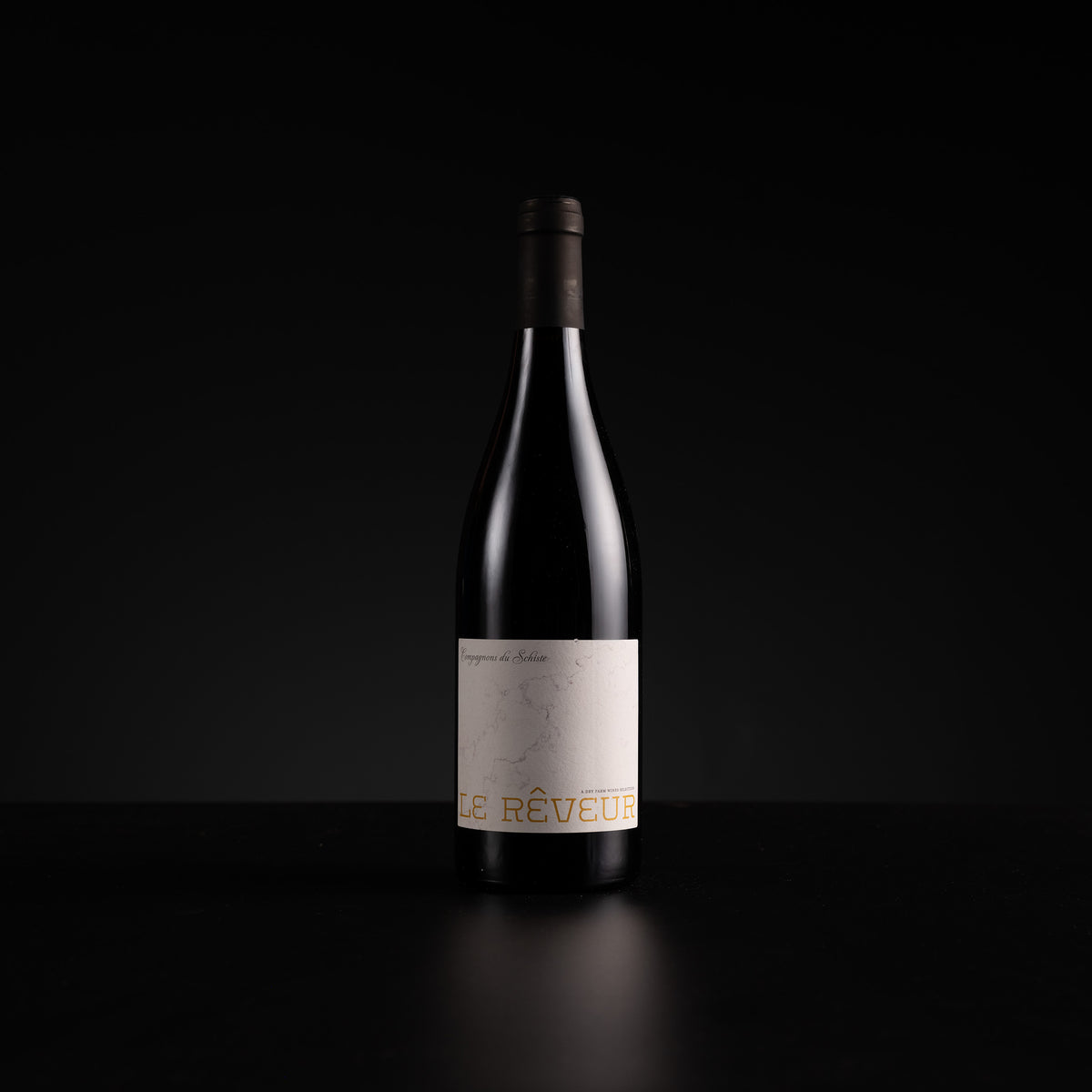 A single dark wine bottle with a minimalist off-white label and gold lettering, standing on a dark reflective surface against a black background.