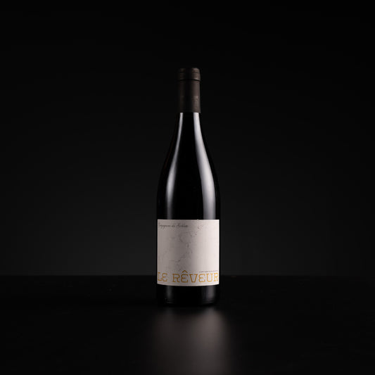 A single dark wine bottle with a minimalist off-white label and gold lettering, standing on a dark reflective surface against a black background.