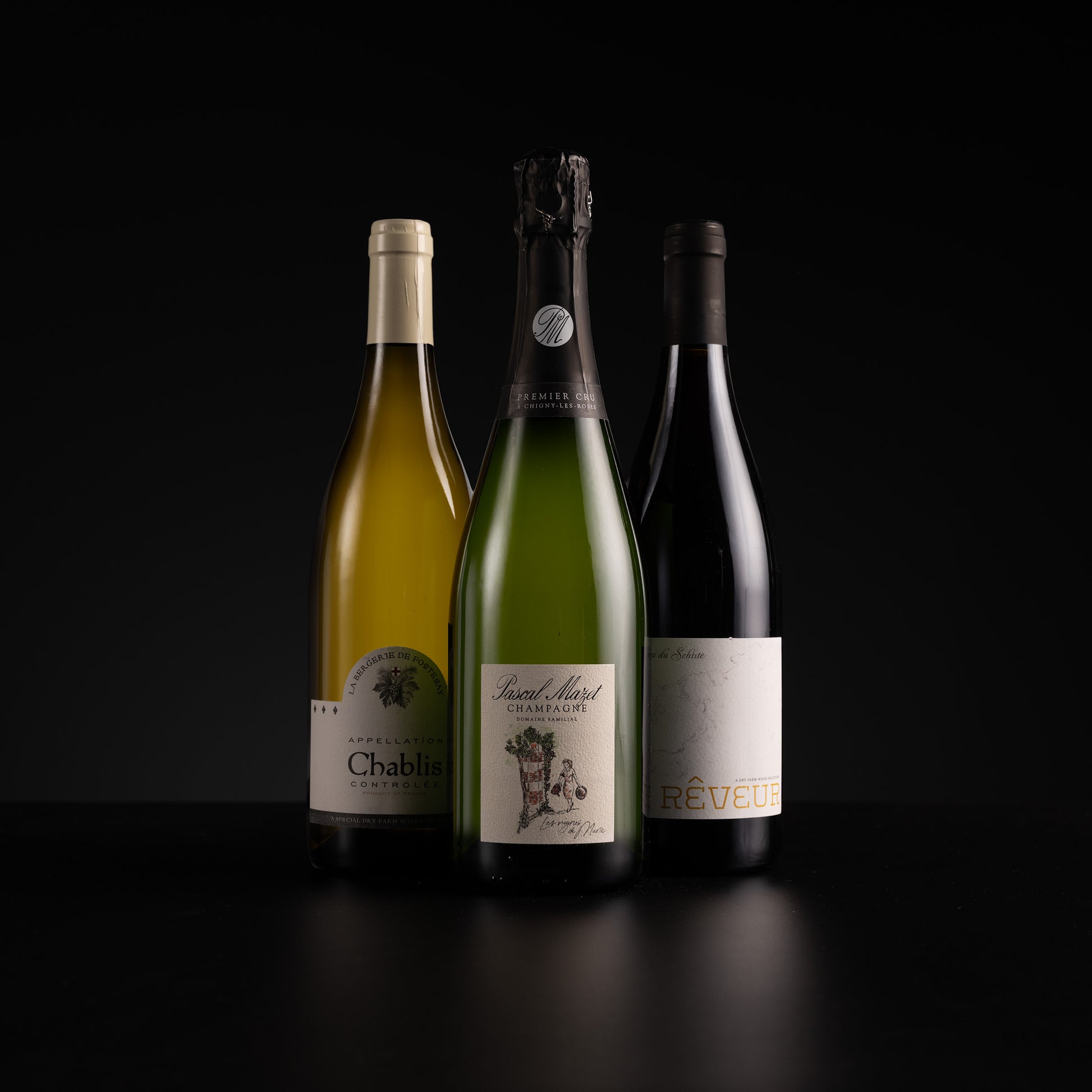 Three wine bottles standing upright against a black background: a pale yellow Chablis bottle on the left, a green sparkling wine bottle in the center, and a dark red wine bottle on the right.