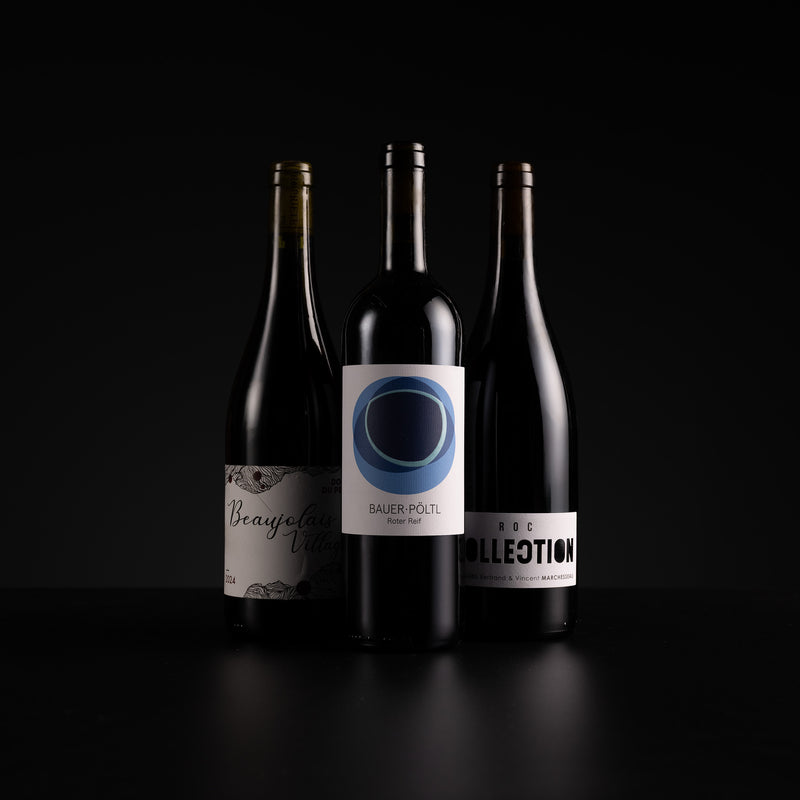 Three dark glass wine bottles standing on a black surface against a black background. The center bottle has a white label with a blue circular design and the text 'BAUER POETZ', while the side bottles have white and gold labels.