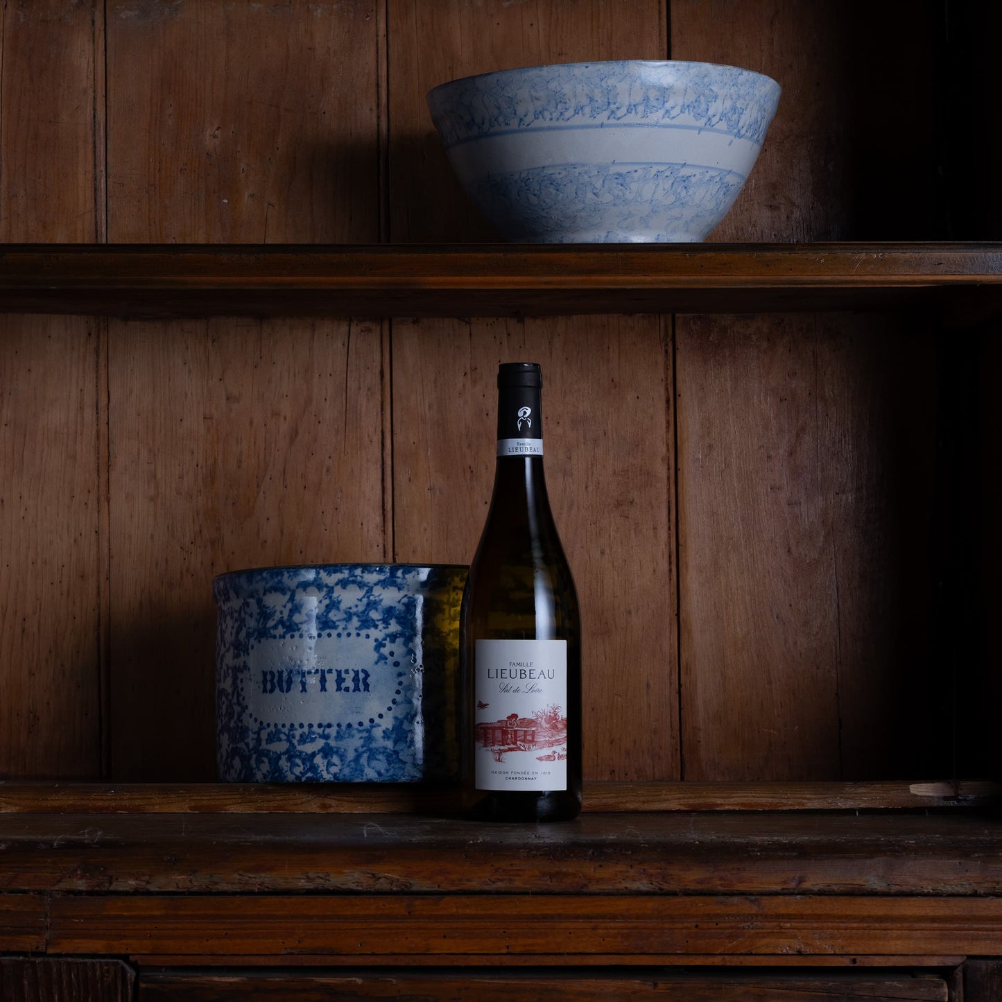 A bottle of La Tour-Rouge Blanc de Blancs wine and a blue-and-white patterned butter tin sit on a rustic wooden shelf next to a matching blue-and-white bowl on a higher shelf, against a dark wood paneled wall.