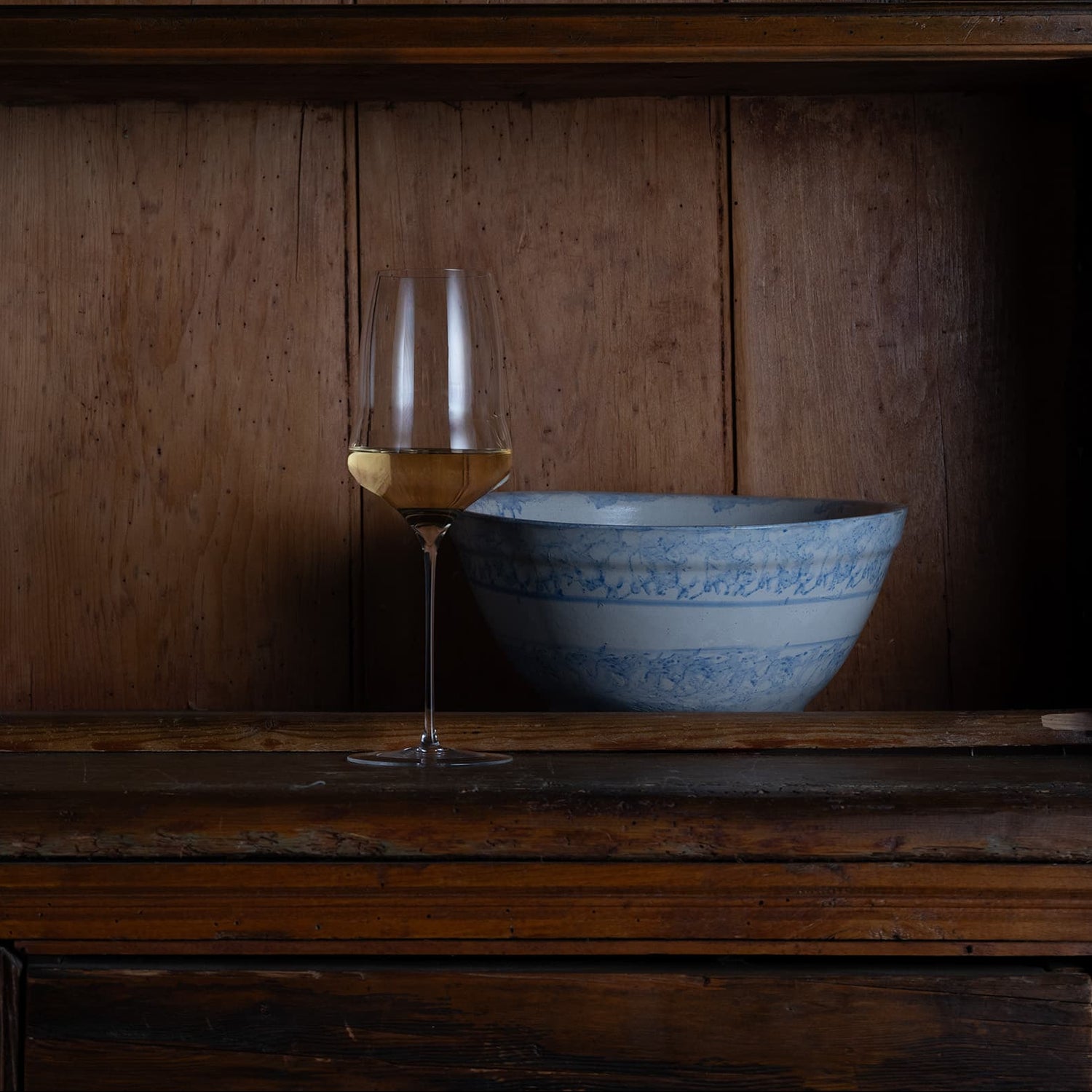 A rustic dark wood hutch with shelves, featuring a light blue and white patterned ceramic bowl and a wine glass on the bottom shelf against a dark background.