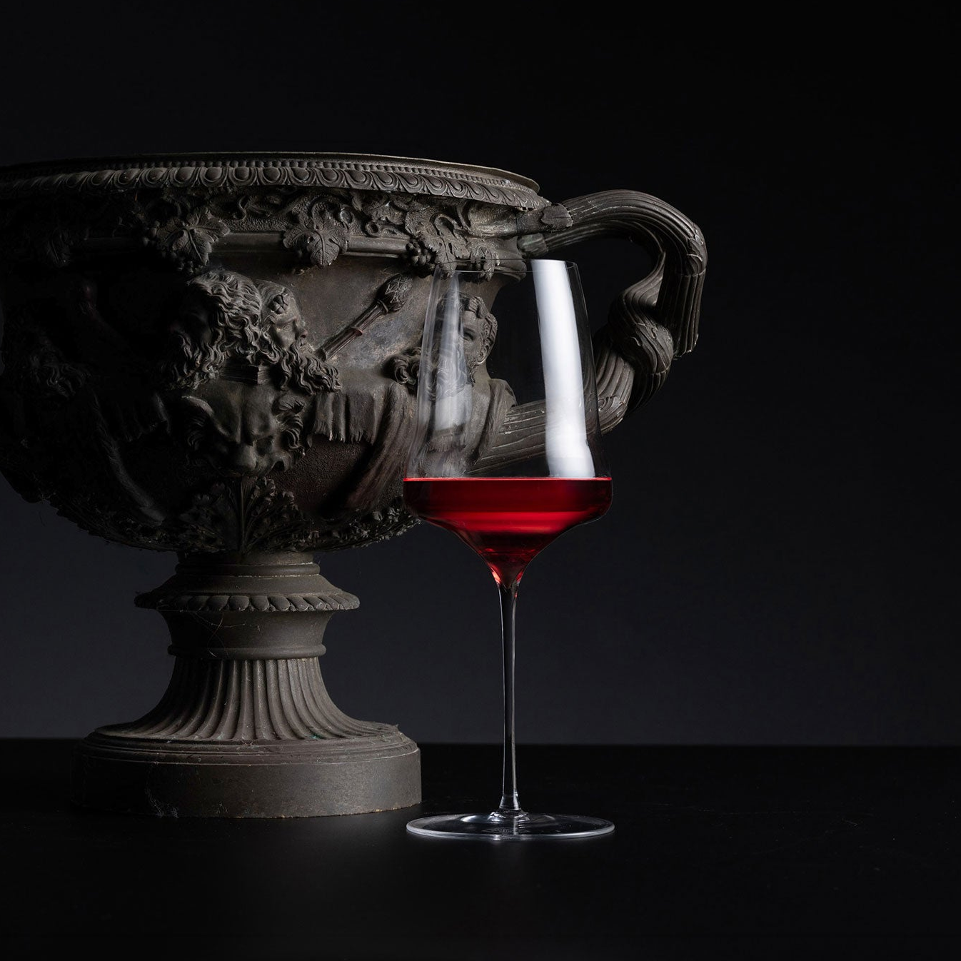 A clear stemmed wine glass partially filled with deep red wine, placed in front of a large ornate dark bronze urn against a black background.