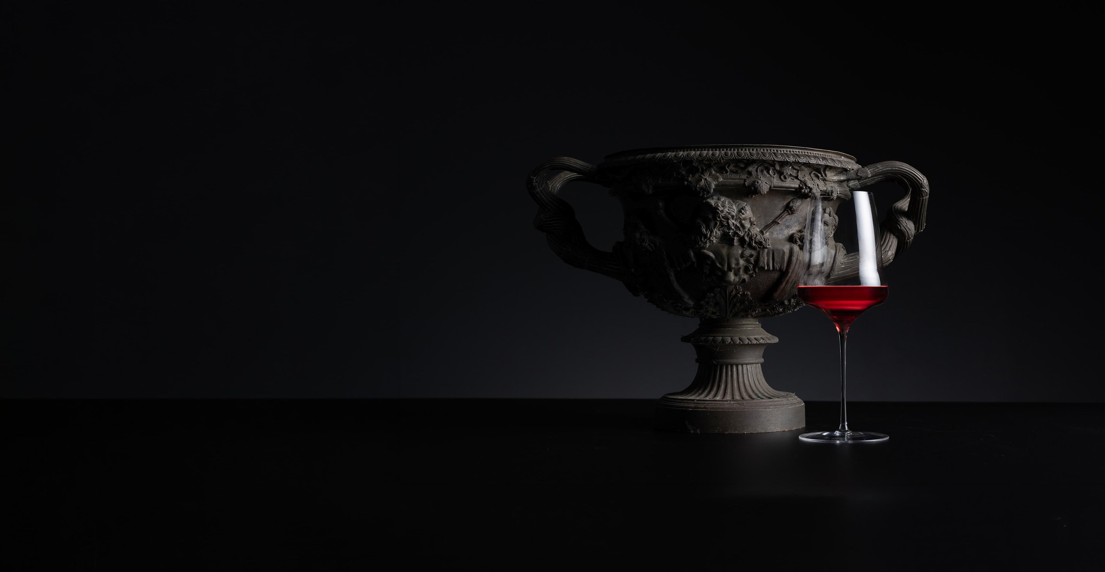 A dark classical-style urn with ornate relief details stands on a black surface against a black background, next to a stemmed glass containing red wine.