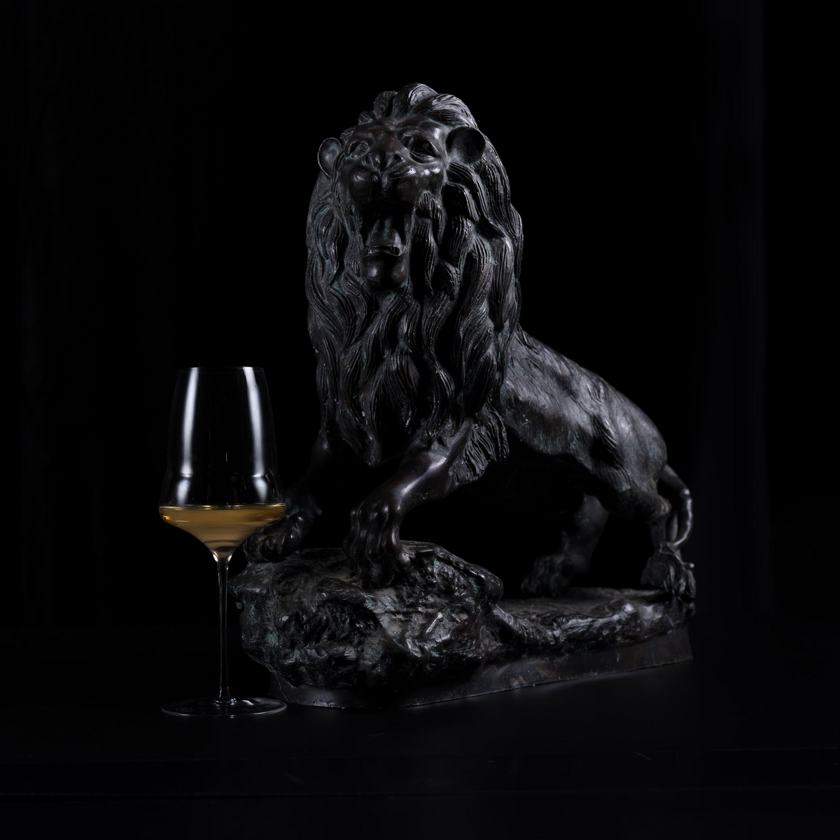 A dark bronze lion sculpture posed on a rocky base, photographed against a black background with a wine glass partially visible at the left side for scale.