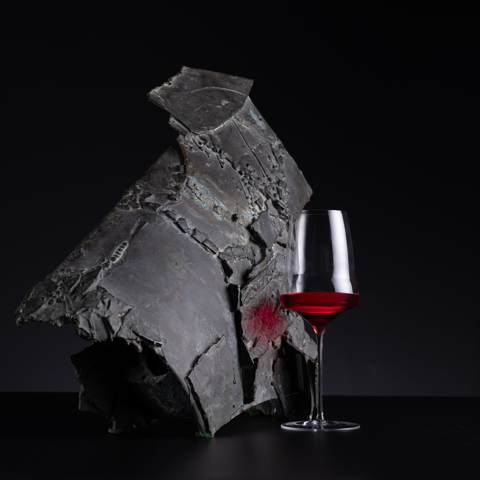 A dark gray, textured, organic-shaped decorative sculpture displayed on a black surface next to a clear wine glass filled with red wine, set against a solid black background.