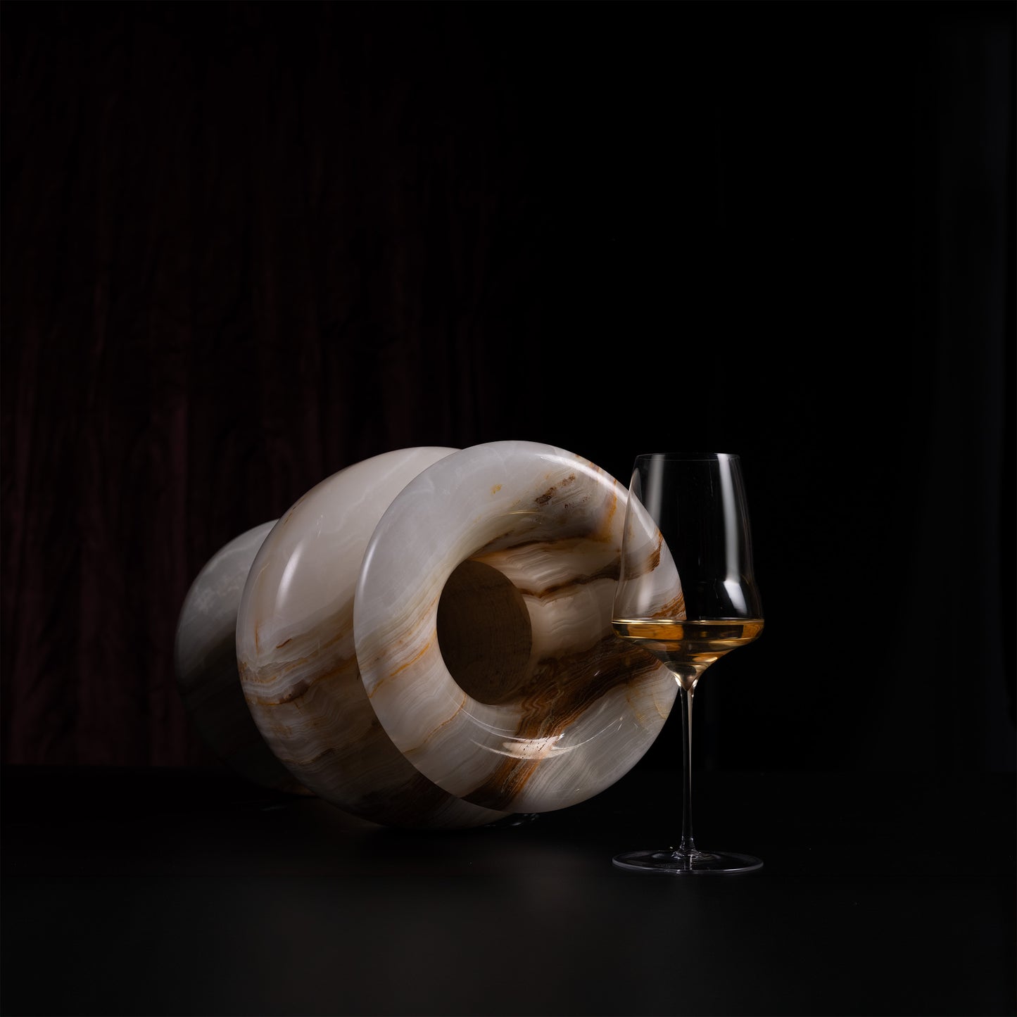 A polished carved stone vessel with a hollow center, shown lying on its side next to a stemmed wine glass containing amber liquid, against a very dark black background.