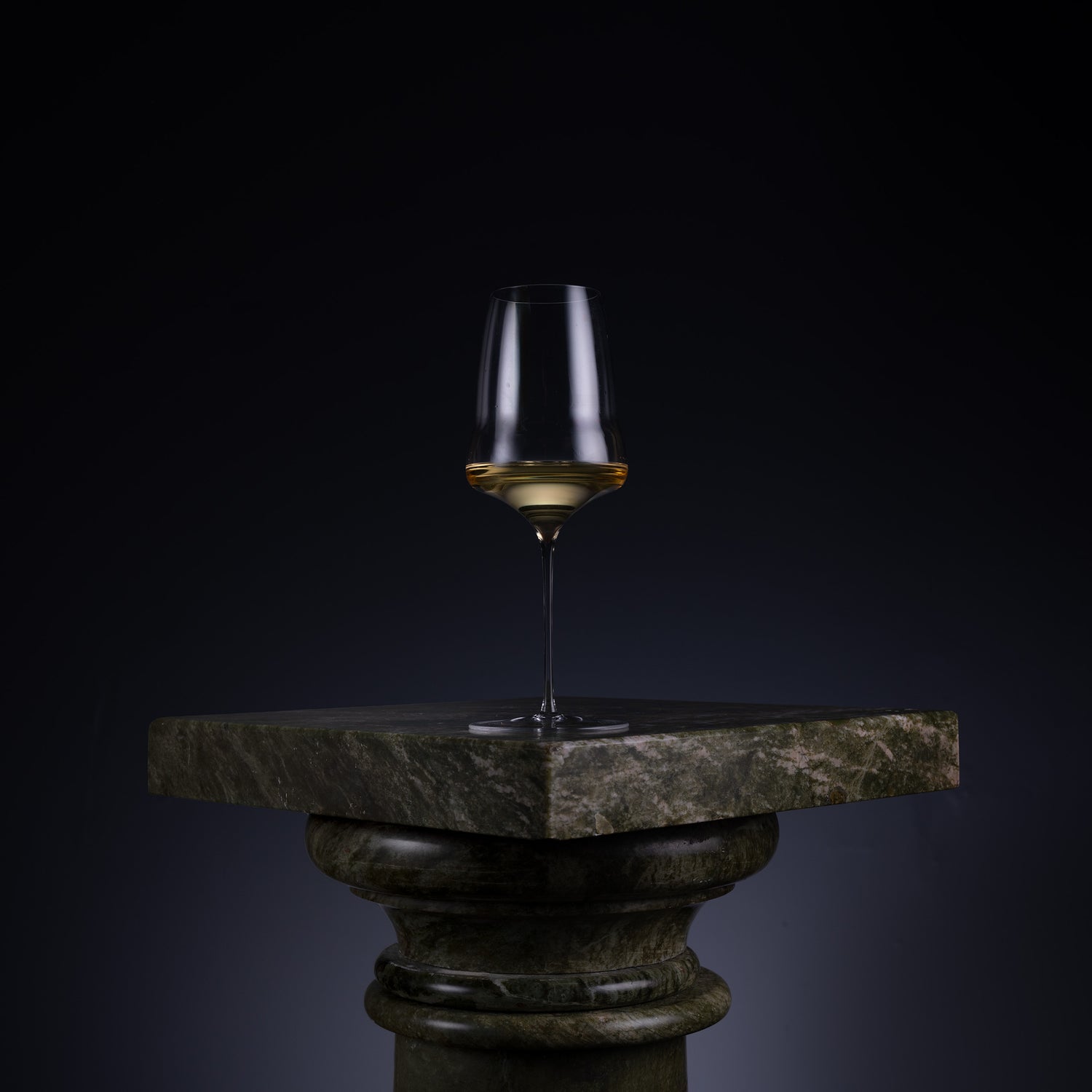 A single stemmed wine glass with a gold band around the base, displayed on a dark stone pedestal against a black background.