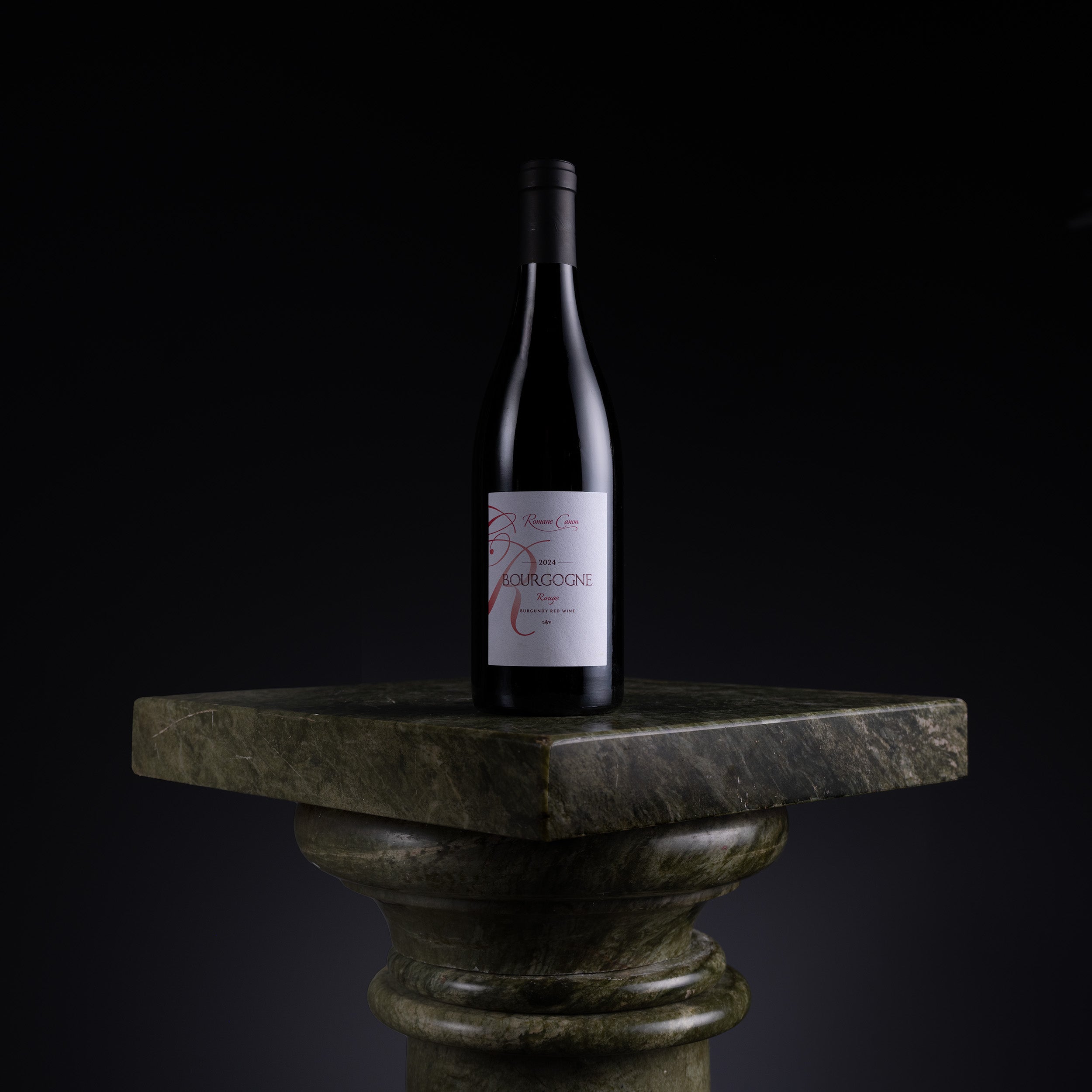 A dark wine bottle with a white label featuring red script, displayed on a dark green stone pedestal against a black background.