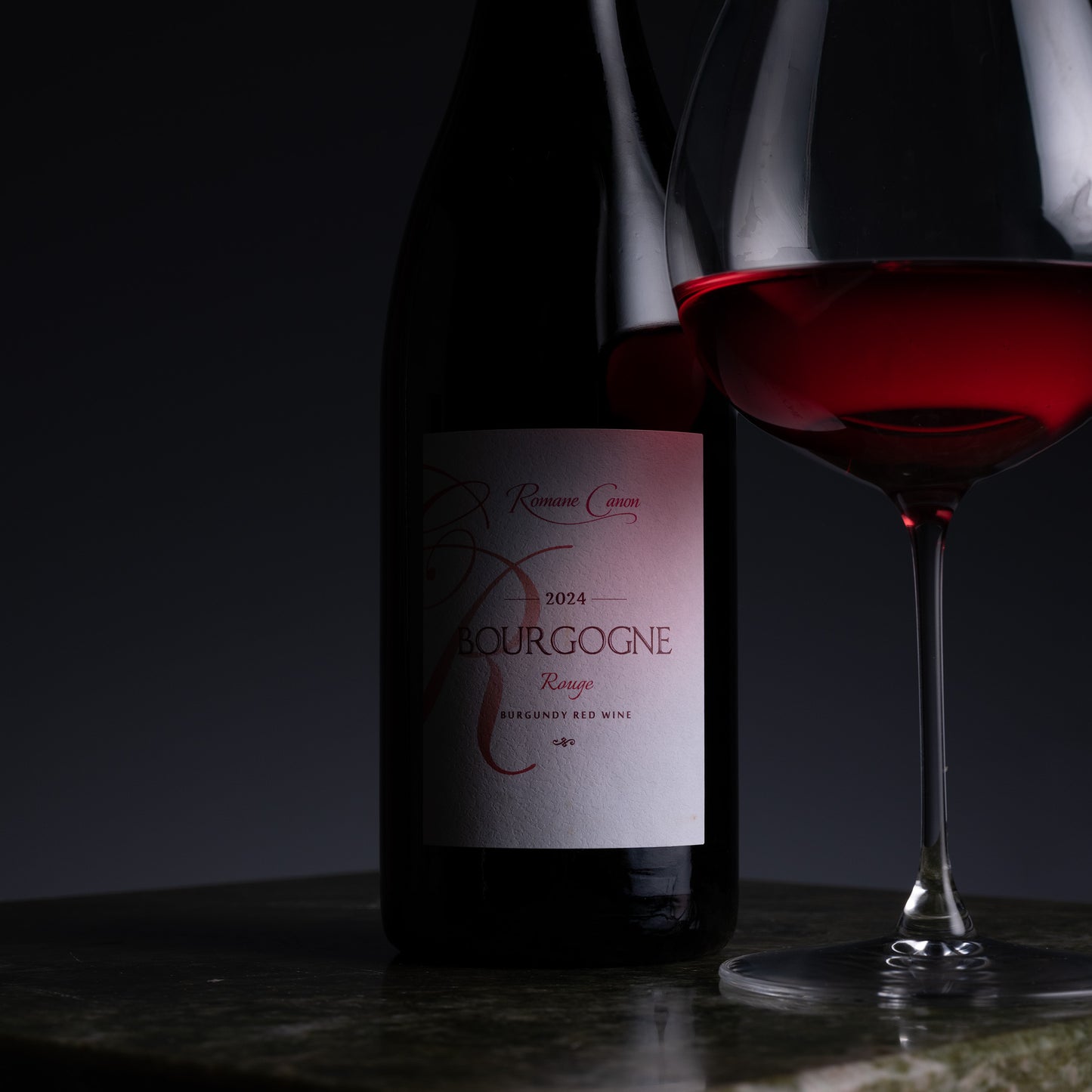 A bottle of Bourgogne Rouge red wine with a glass of the deep red wine poured next to it, set on a dark reflective surface against a dark background.