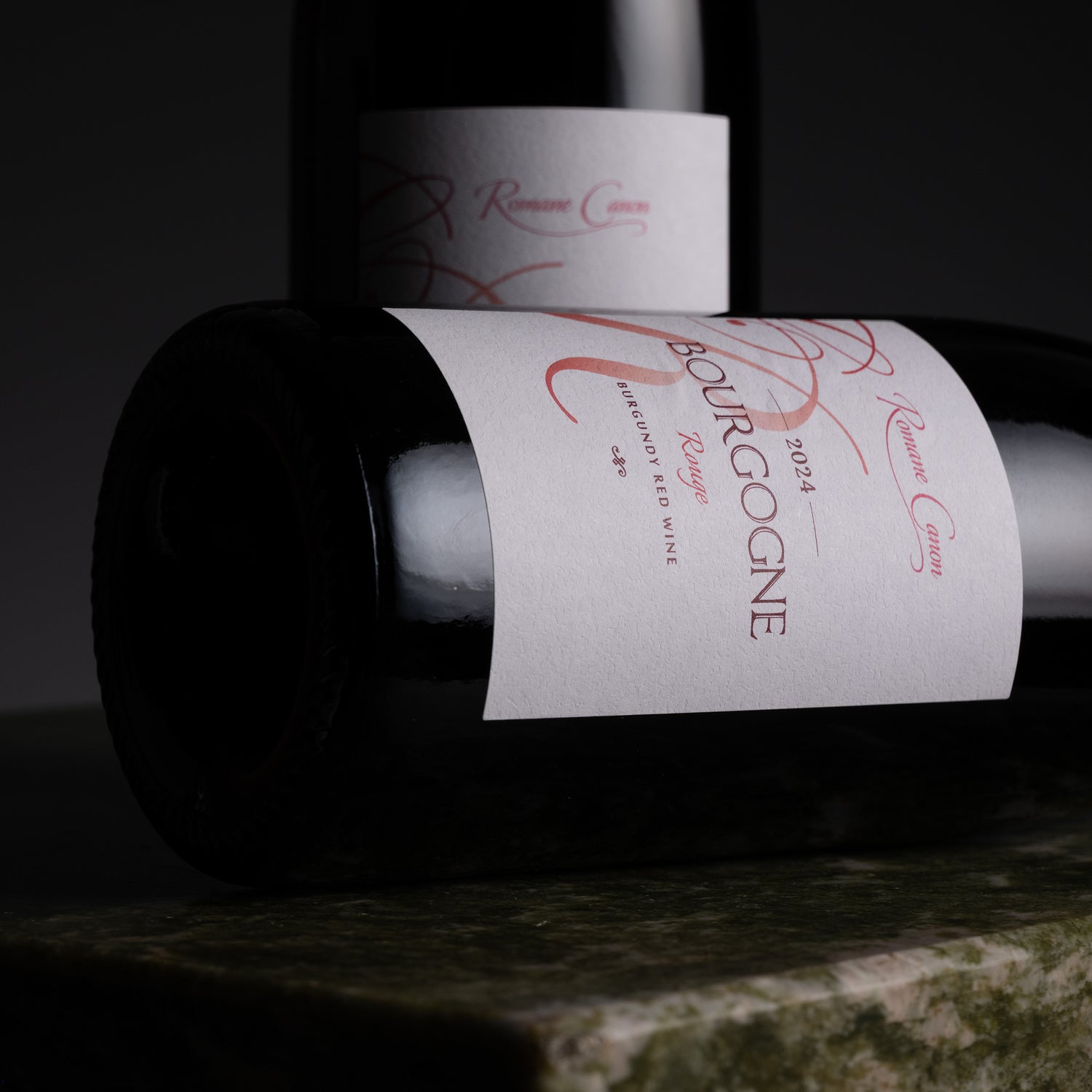 A close-up of a dark wine bottle lying horizontally with a white paper label reading 'Bourgogne' and 'Riviera Costa' in red script, set against a dark background with another bottle blurred behind it.