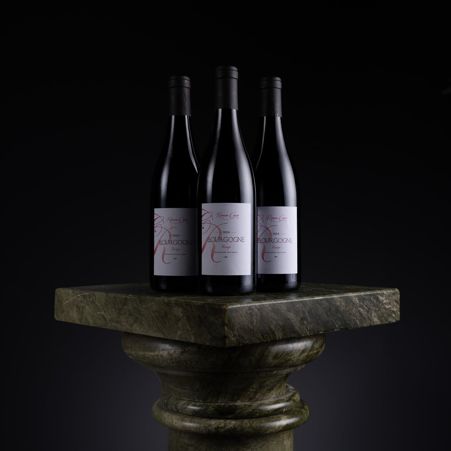 Three dark wine bottles with white labels featuring red script text, displayed on a green stone pedestal against a black background.