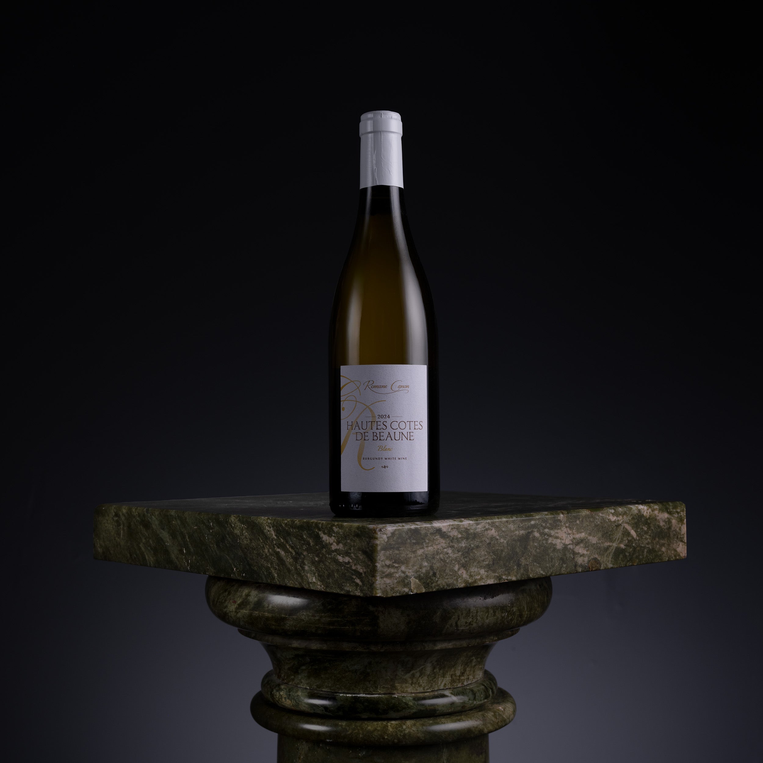A single wine bottle with a white label stands centered on a dark stone pedestal against a black background. The bottle appears olive-brown glass with a white capsule and a white label featuring dark text.