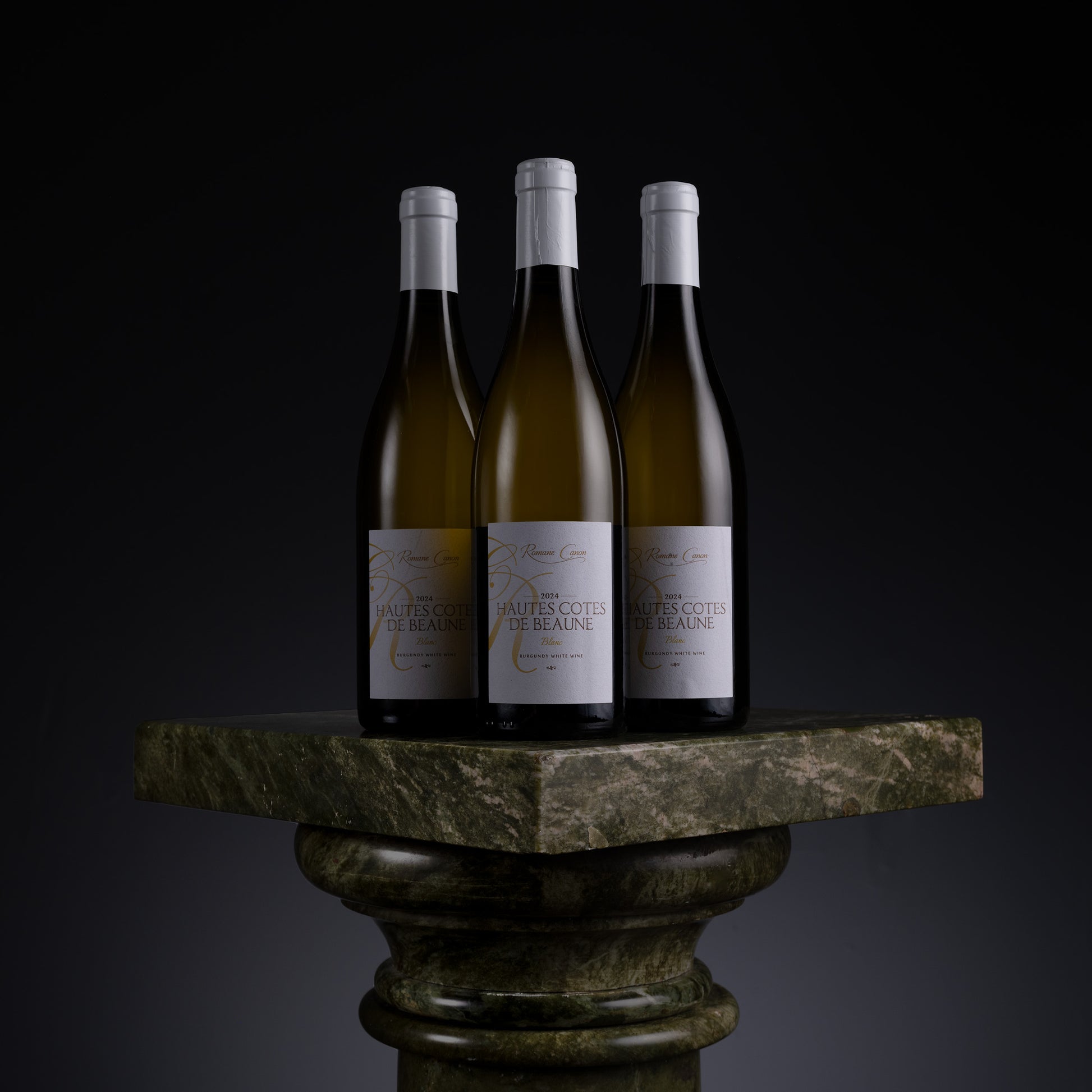 Three bottles of white wine displayed side by side on a dark green marble pedestal against a black background. The labels read 'Maitre Coeur de Vain' and 'Saint-Ouen'.