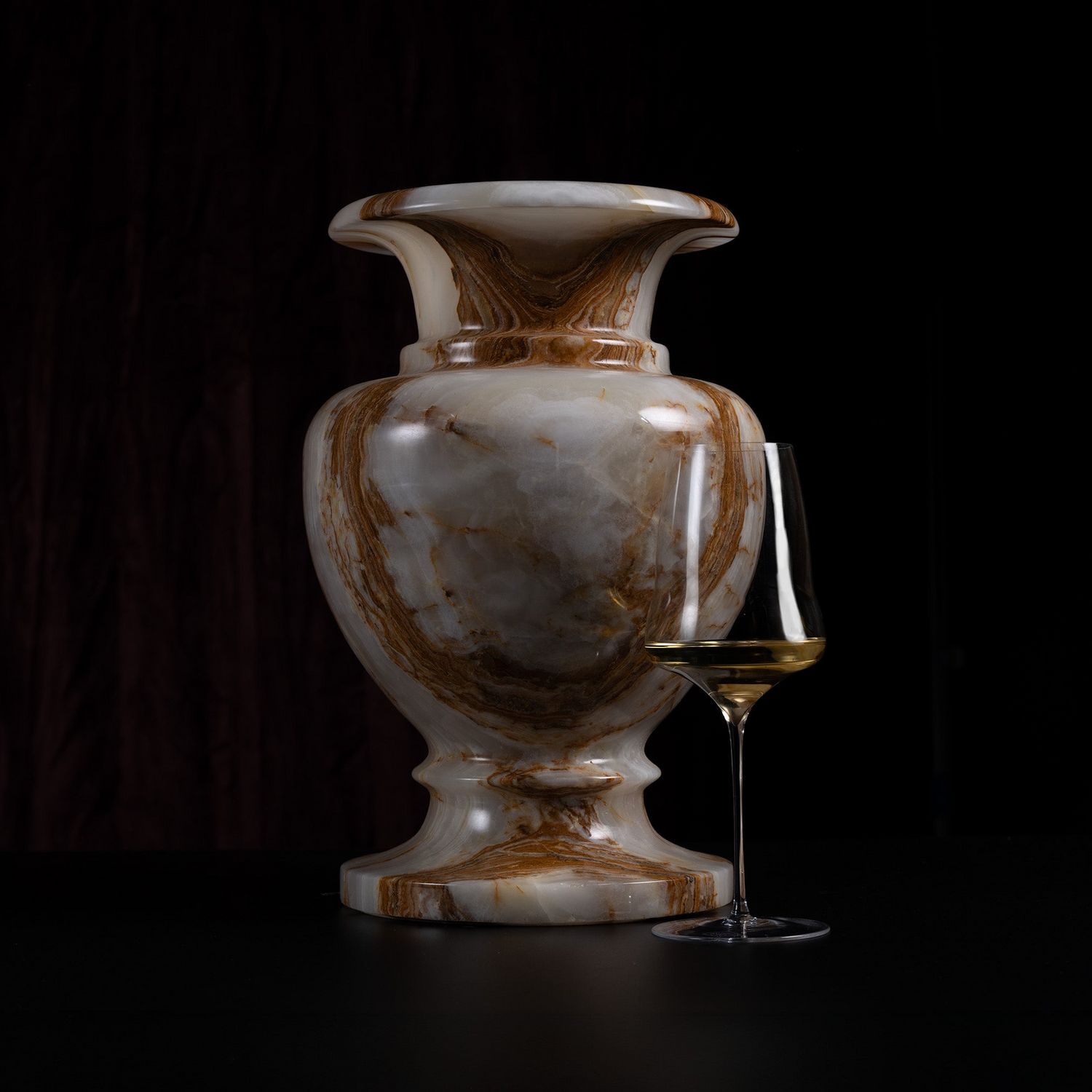 A polished onyx marble vase with swirling brown and cream patterns, shown next to a wine glass on a dark background.
