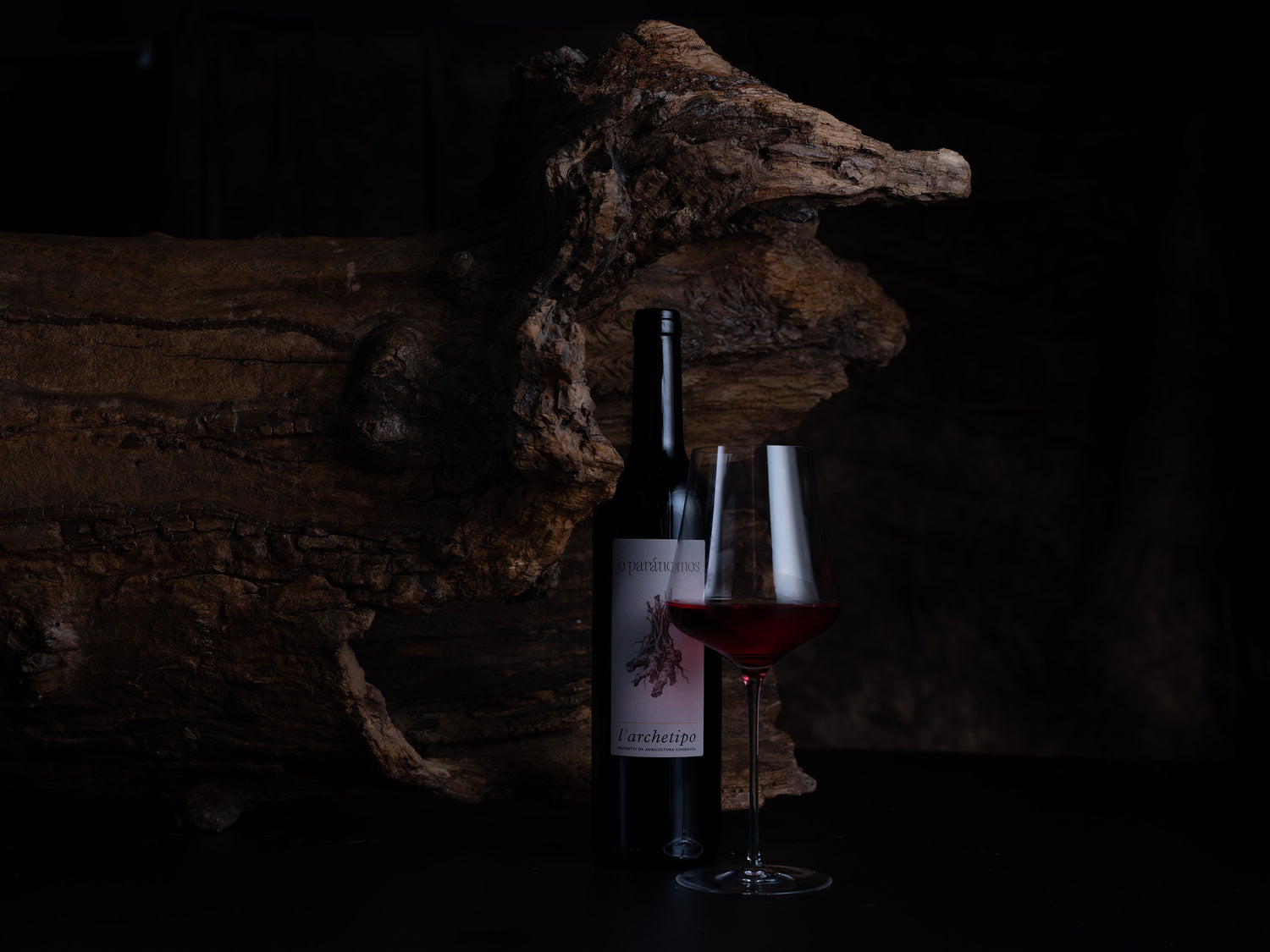 A dark wine bottle with a partially visible label stands beside a stemmed wine glass filled with red wine, set against a very dark background with a large piece of driftwood.