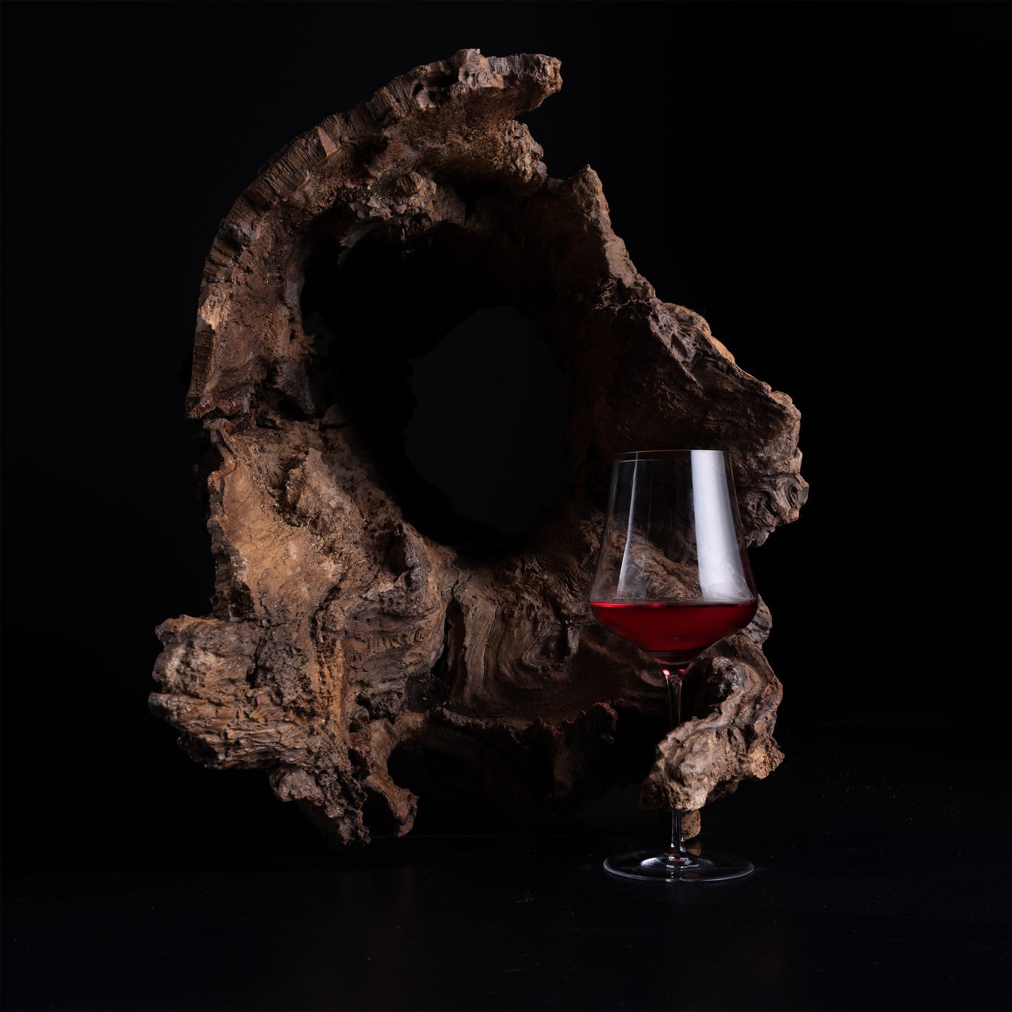 A clear wine glass partially filled with red wine, placed in front of a large natural wood driftwood piece on a black background.