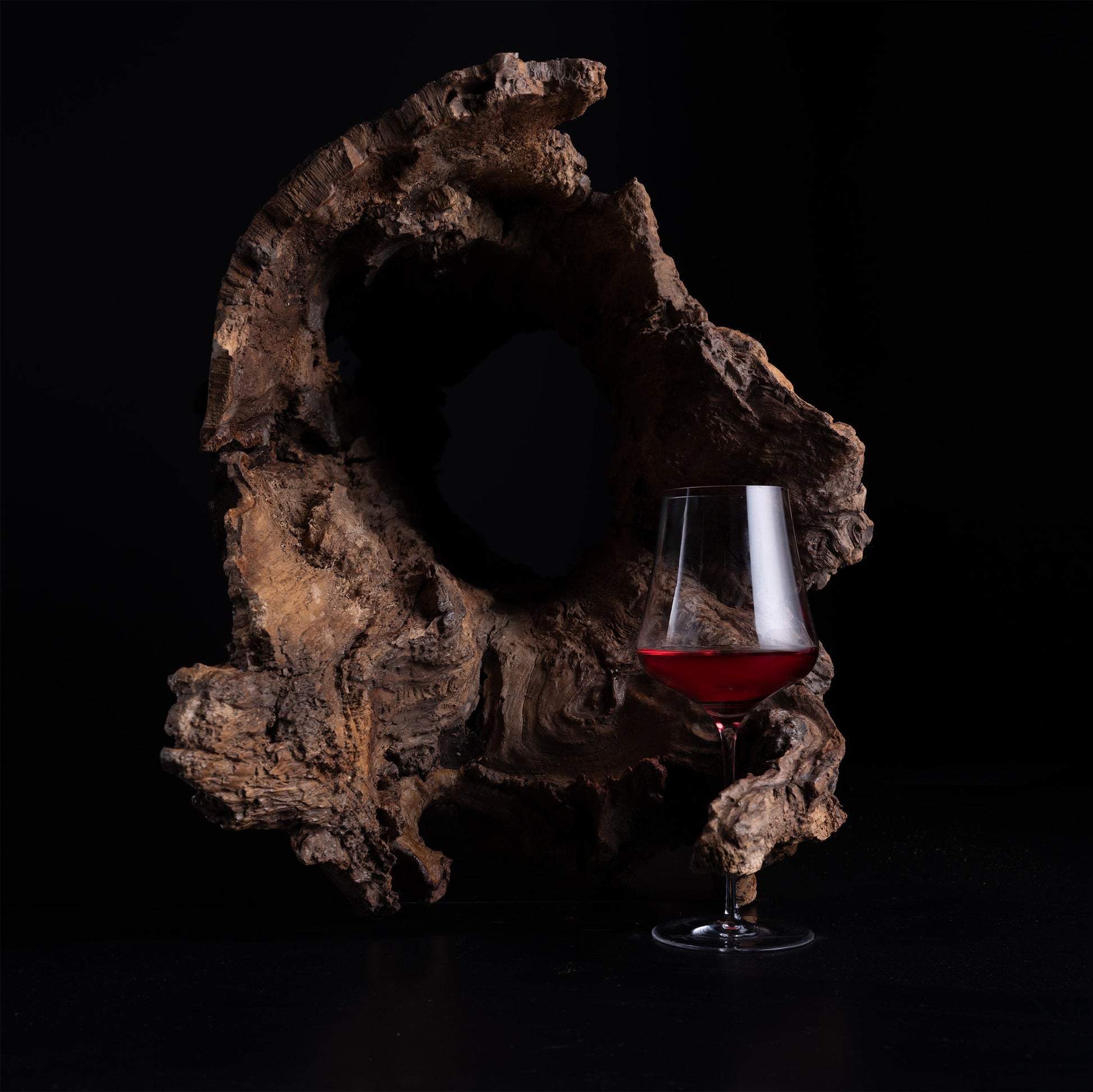 A clear wine glass partially filled with red wine, placed in front of a large natural wood driftwood piece on a black background.