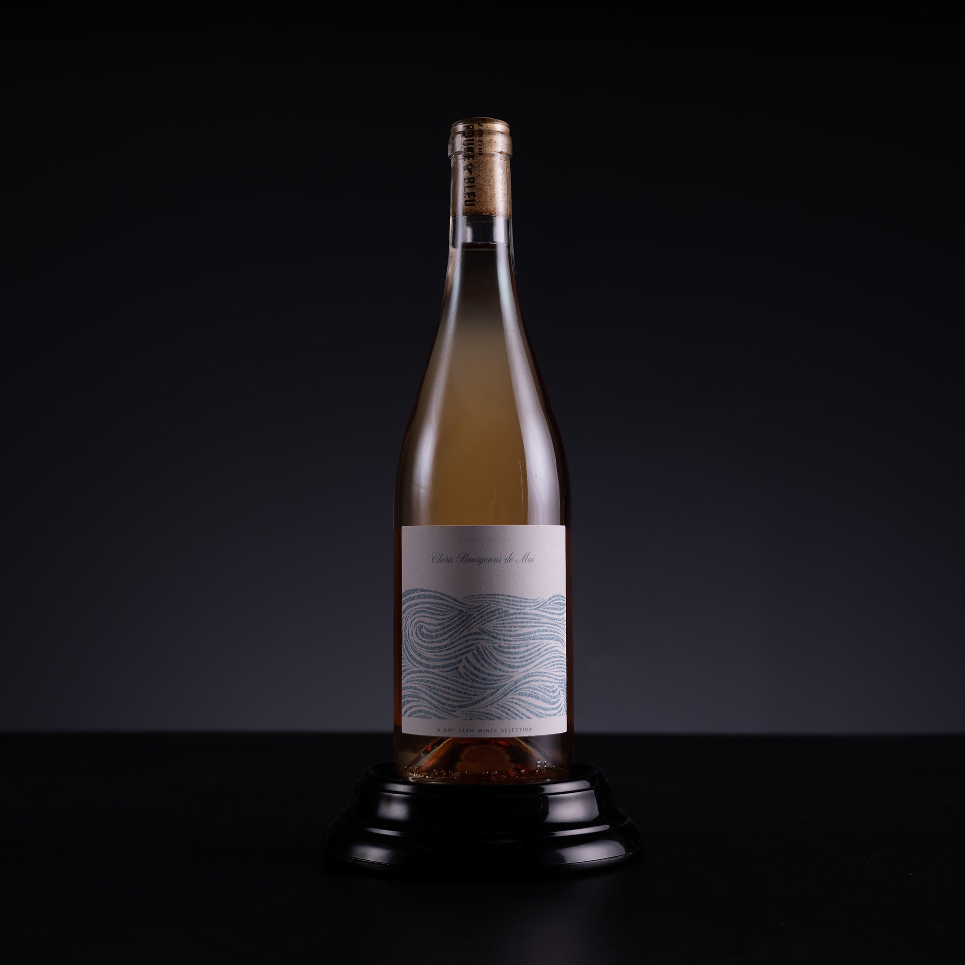 A single wine bottle with a gold cap and a white label featuring a blue wave pattern, displayed on a black stand against a dark studio background.