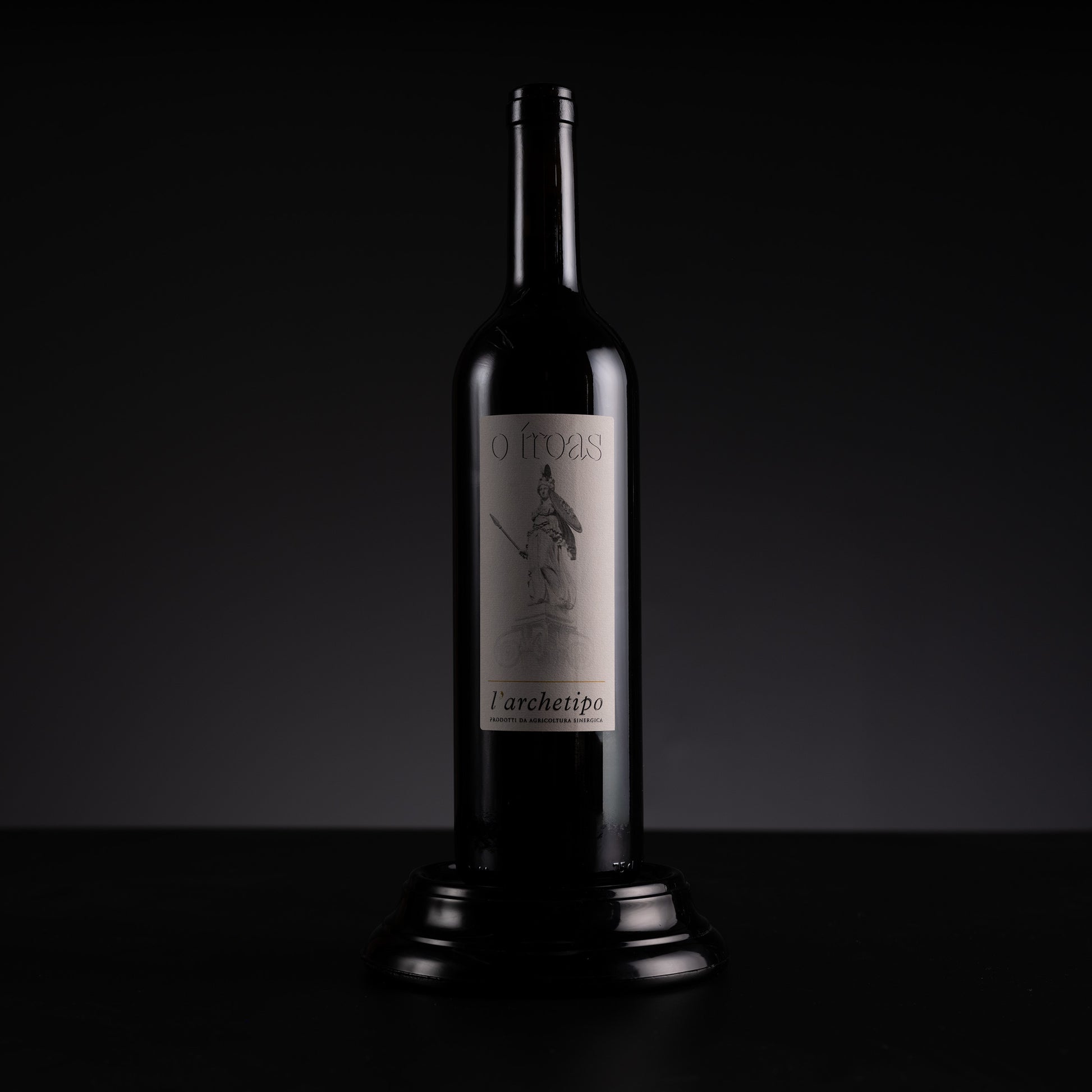 A dark glass wine bottle with a silver label reading 'O Troas' and 'l'architetto', displayed on a black pedestal against a dark studio background.
