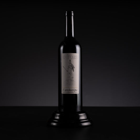 A dark glass wine bottle with a silver label reading 'O Troas' and 'l'architetto', displayed on a black pedestal against a dark studio background.