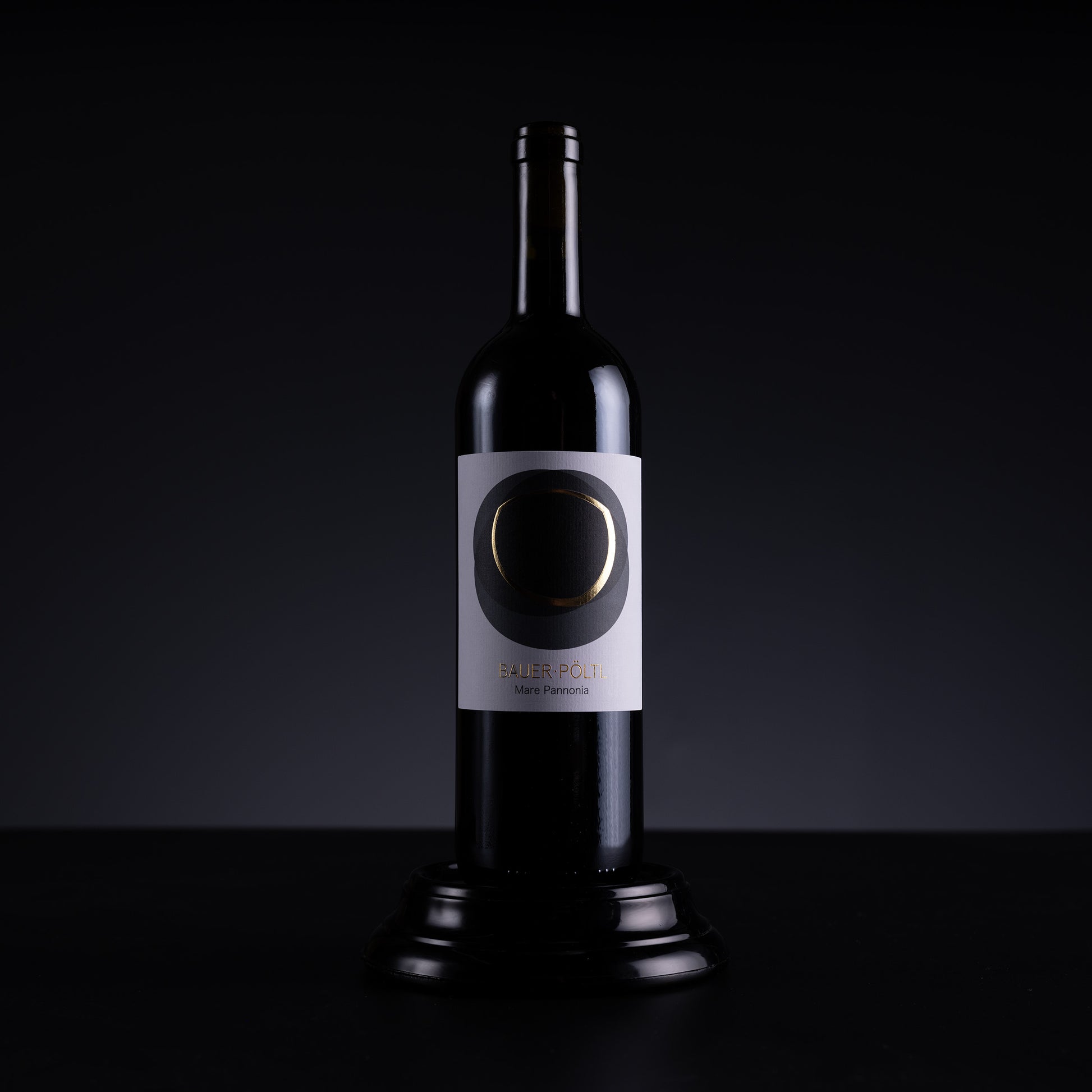A dark glass wine bottle with a white label featuring a circular black and gold design, displayed on a black pedestal against a dark studio background.