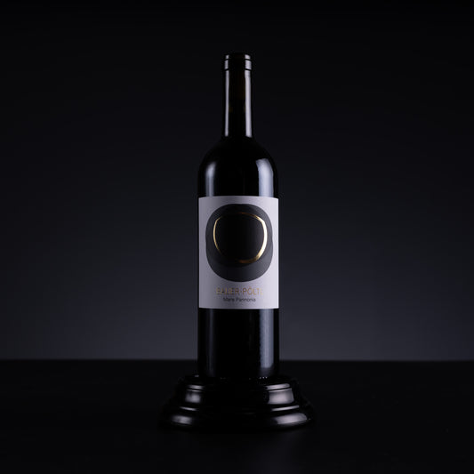A dark glass wine bottle with a white label featuring a circular black and gold design, displayed on a black pedestal against a dark studio background.