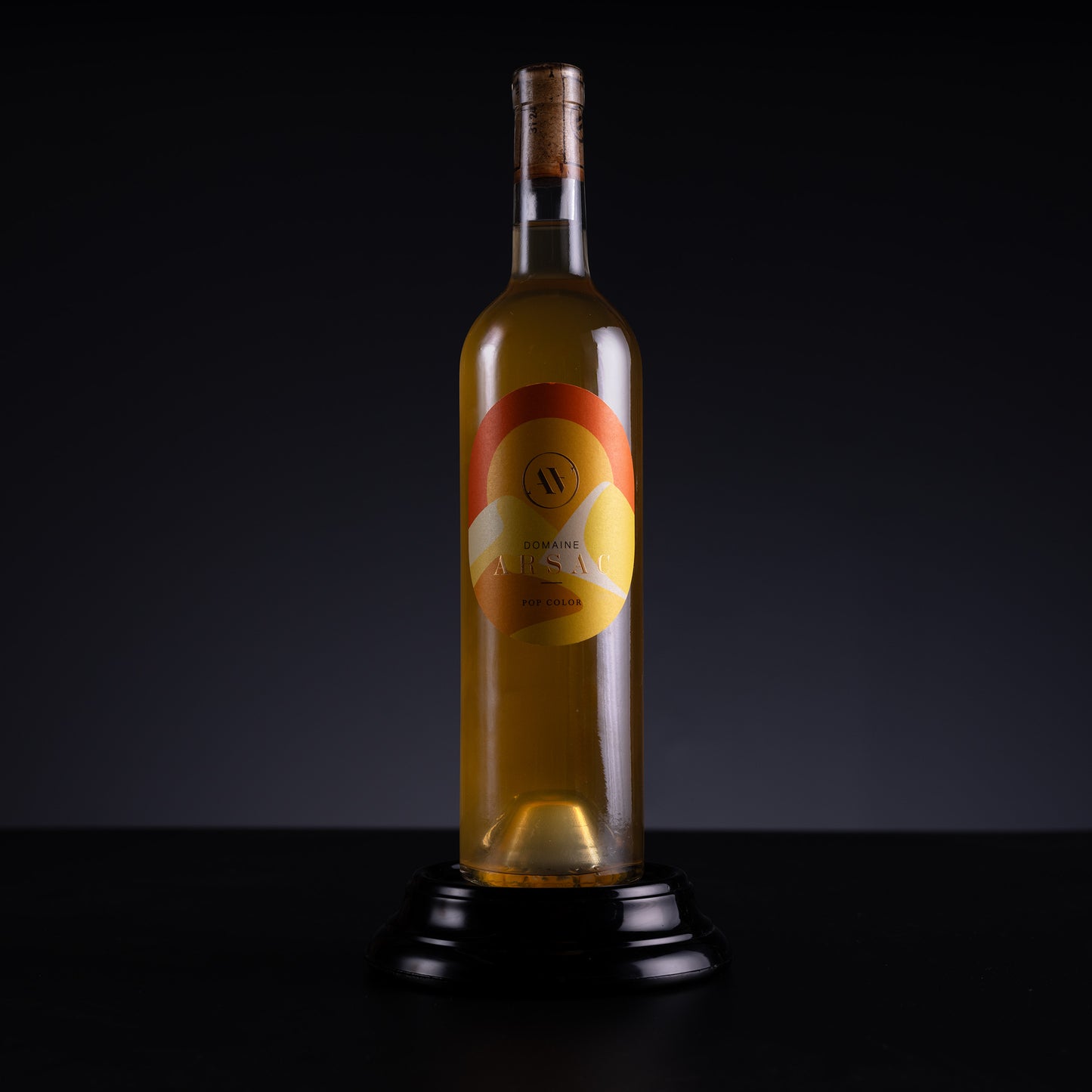 A tall glass bottle filled with golden amber liquid, standing upright on a black pedestal against a dark studio background. The bottle has a cork stopper and a round label with orange, yellow, and white graphics.