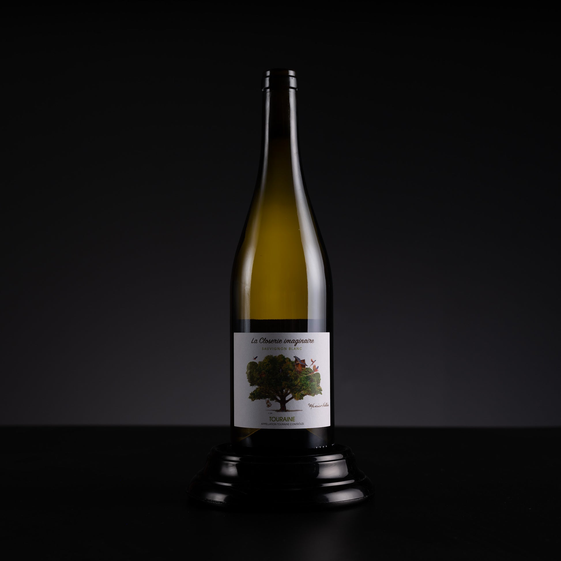 A single wine bottle with a white label featuring a tree illustration, displayed on a black stand against a dark studio background.