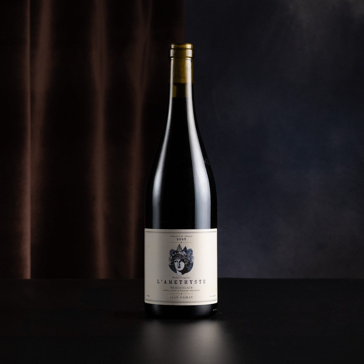A single dark wine bottle with a cream label stands centered on a dark surface against a moody black and brown backdrop.