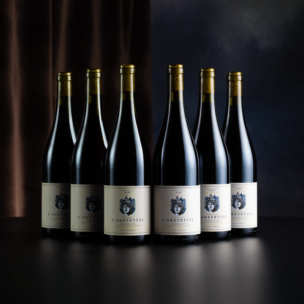 Six dark wine bottles with cream labels and gold foil caps, arranged in a row on a dark surface against a dark draped background. The labels show a crest and the text 'L'ANCIENNE'.