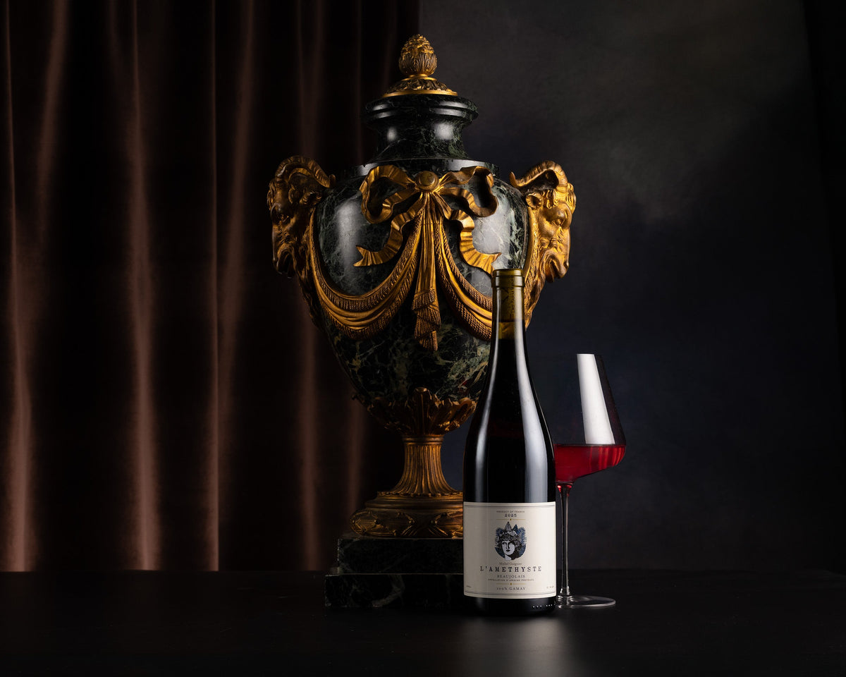 A bottle of red wine with a white label stands next to a filled wine glass, set against an ornate black and gold urn and dark draped curtains.