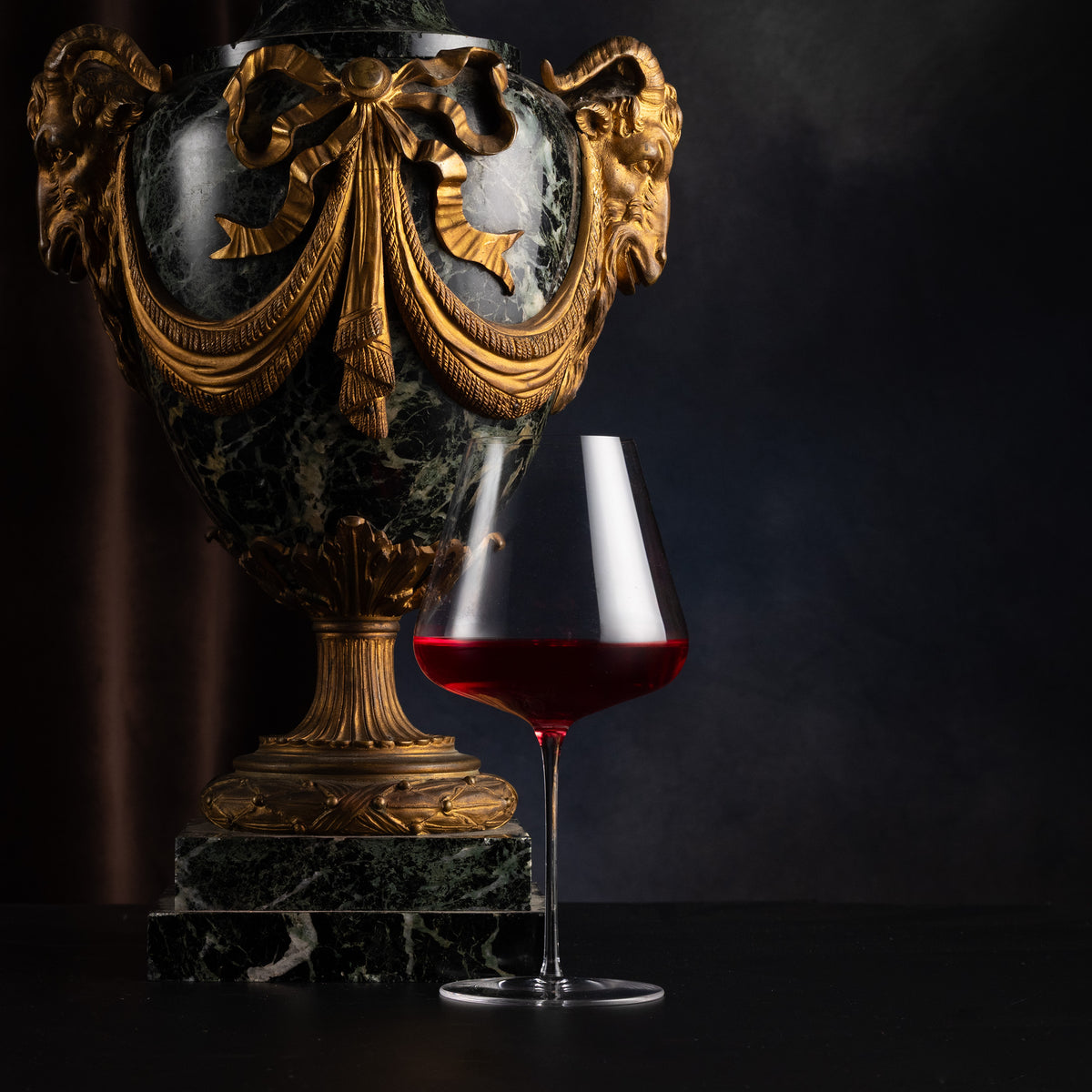 An ornate dark green marble and gold decorative urn displayed next to a stemmed wine glass filled with red wine on a dark background.
