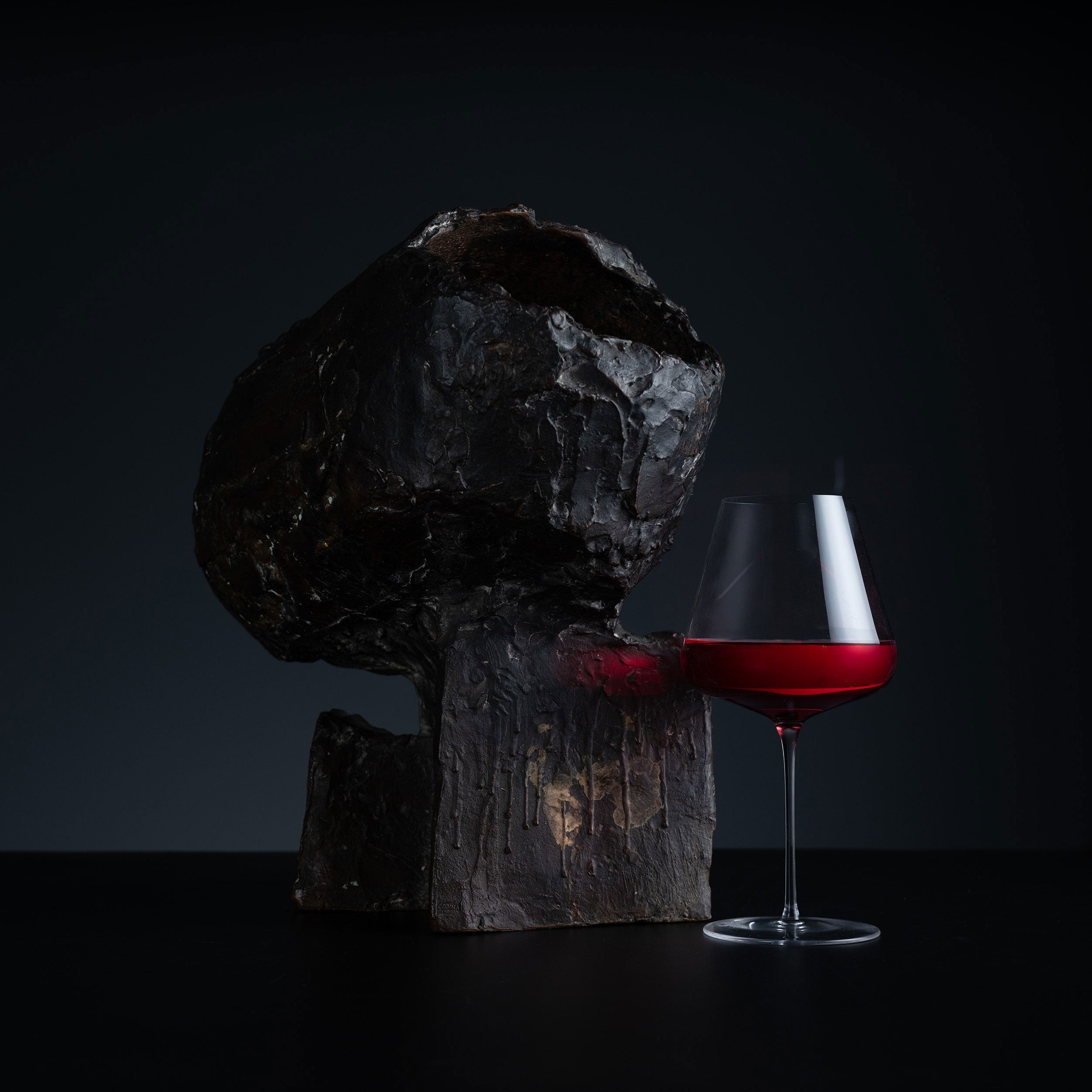A dark, rustic wooden sculpture or decorative block beside a stemmed wine glass filled with red wine, set against a very dark studio background.